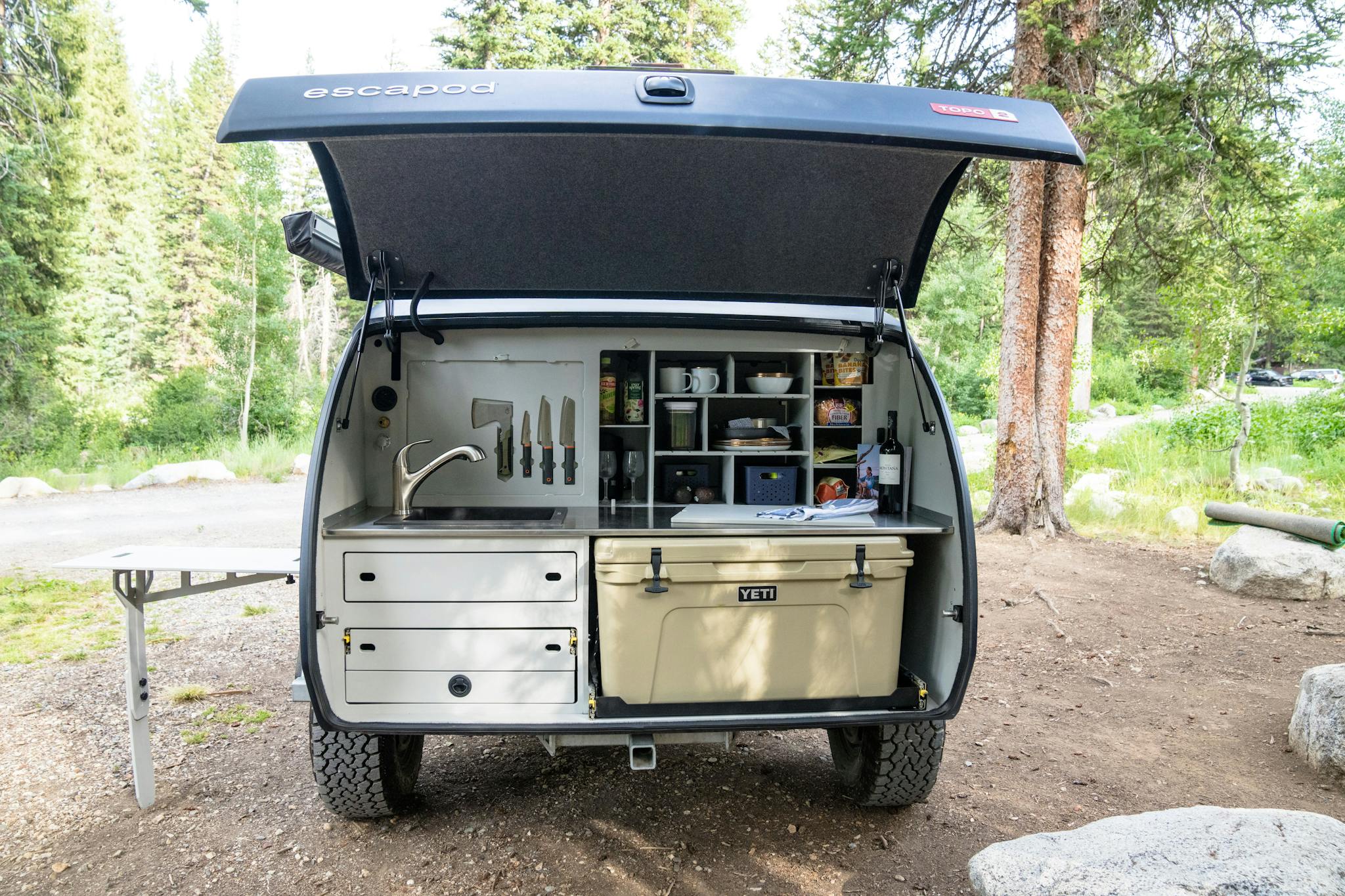 A teardrop trailer with a full kitchen set-up. A sink, YETI cooler, storage, and two-burner stove.