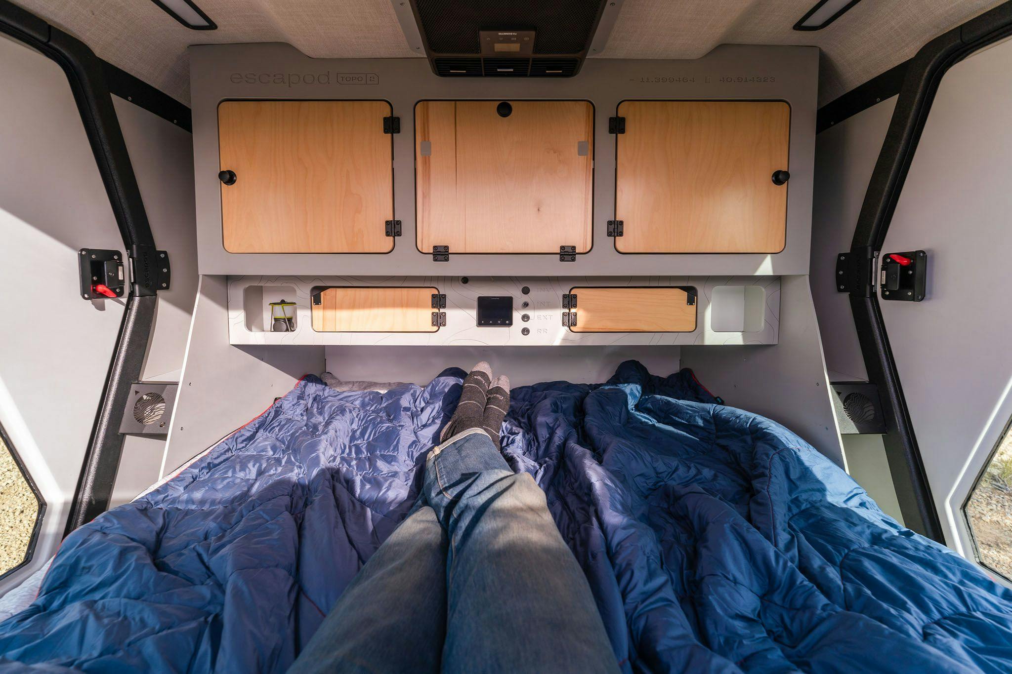 A man is lounging in the spacious cabin of the TOPO2 teardrop camper with his legs extended and crossed at the ankles.