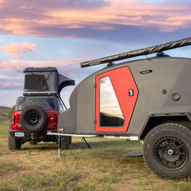 A TOPO2, a teardrop trailer, with a mounted awning parked beside a red Bronco with a rooftop tent mounted on top.
