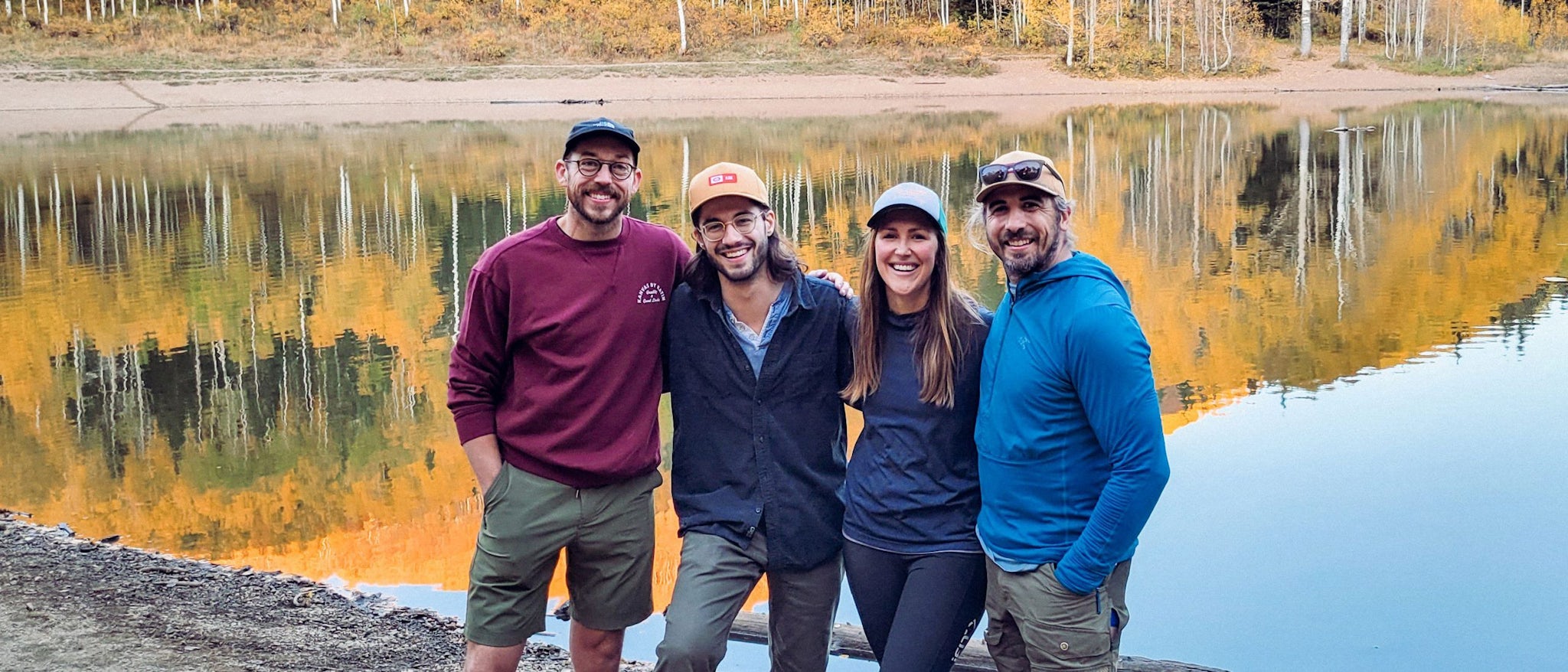 Four people smiling in front of a lake front with the reflection of fall leaves.