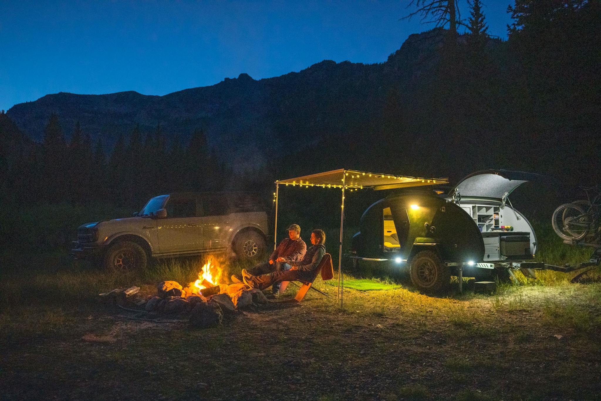 A couple sit around the fire while their TOPO2, a custom offroading trailer, sits  glow behind them.