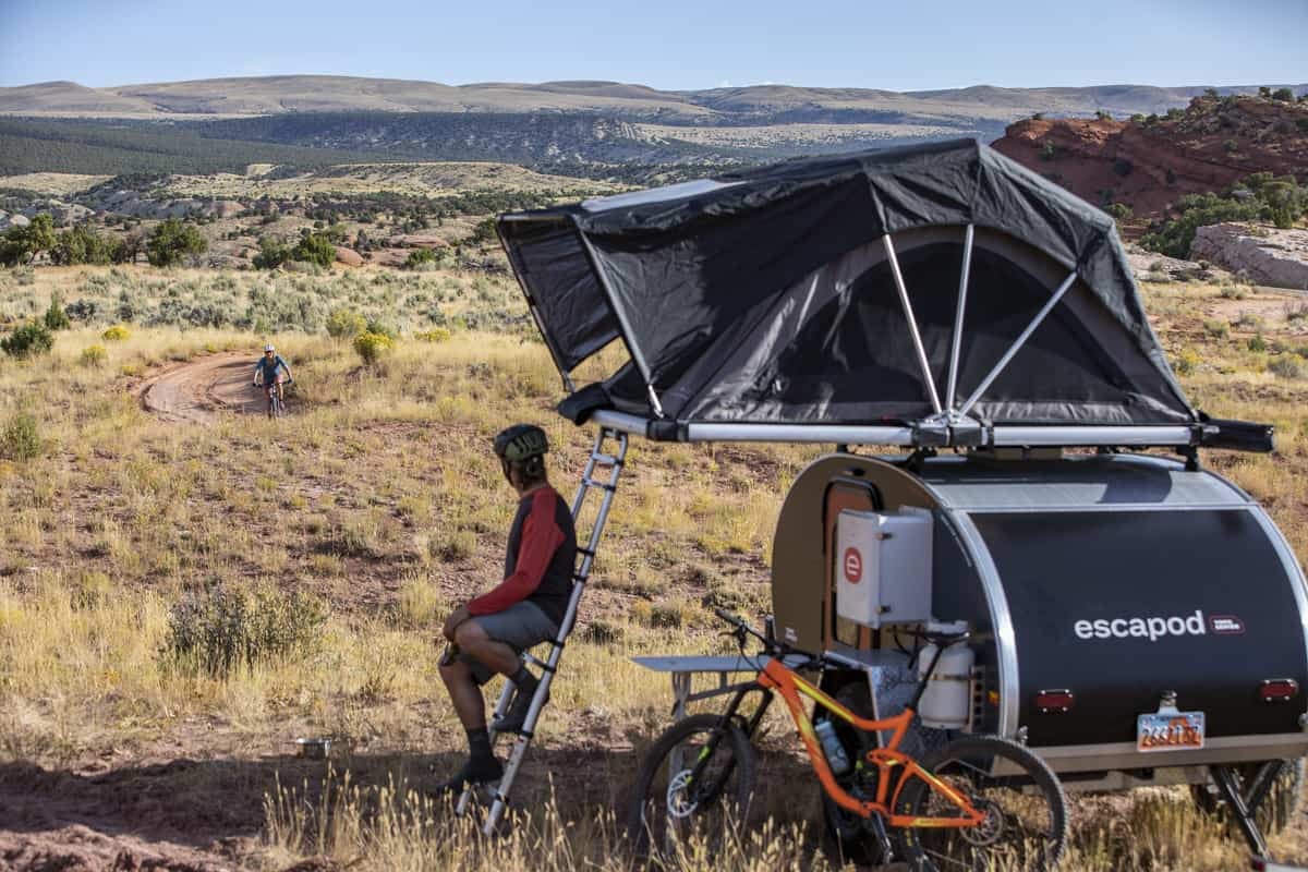 A man resting against his teardrop camper with a rooftop tent mounted.