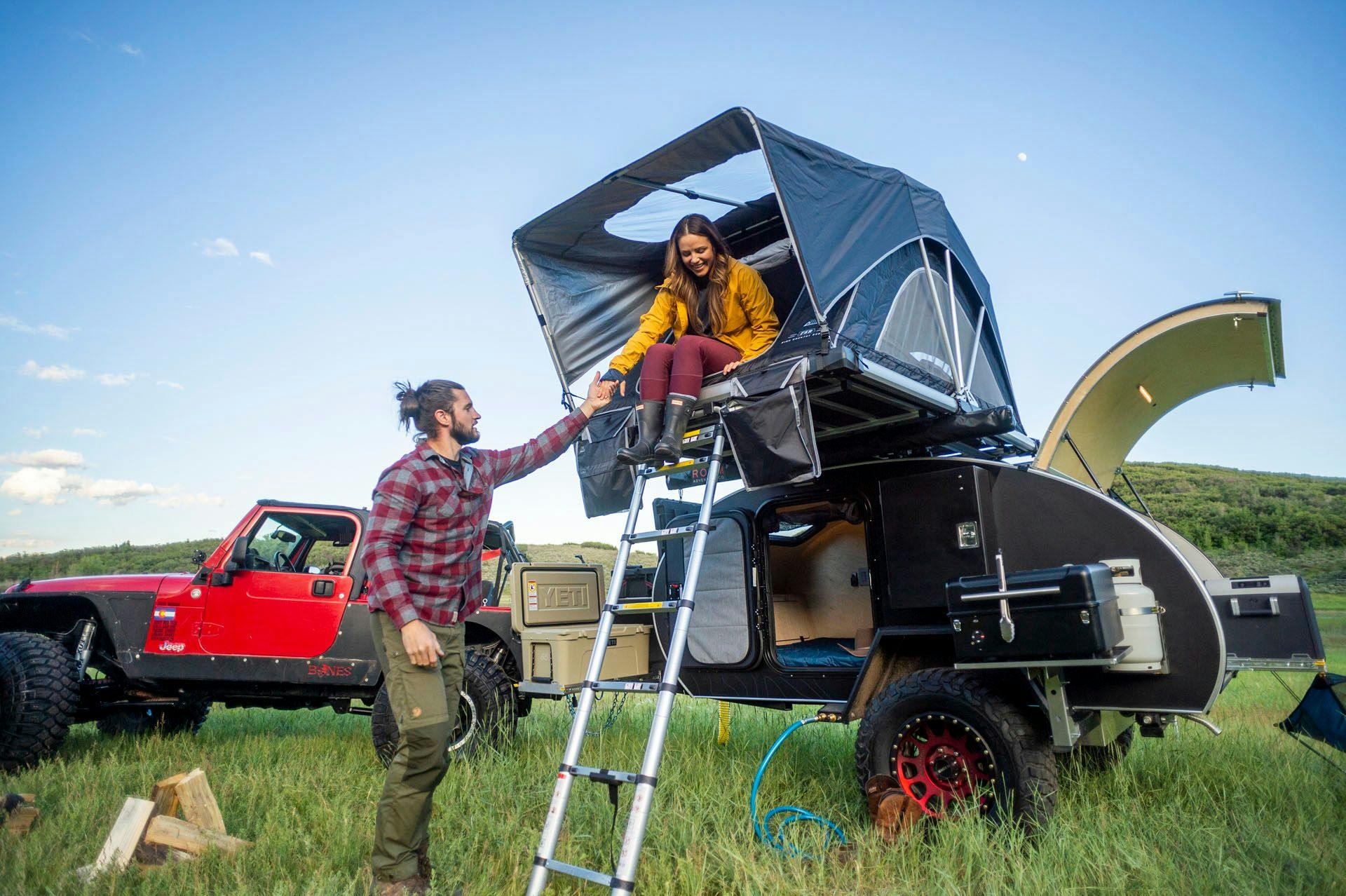 A man in a red plaid shirt, helps a woman in a yellow rain jacket, down the ladder of a rooftop tent on top of an Escapod Original TOPO teardrop trailer.