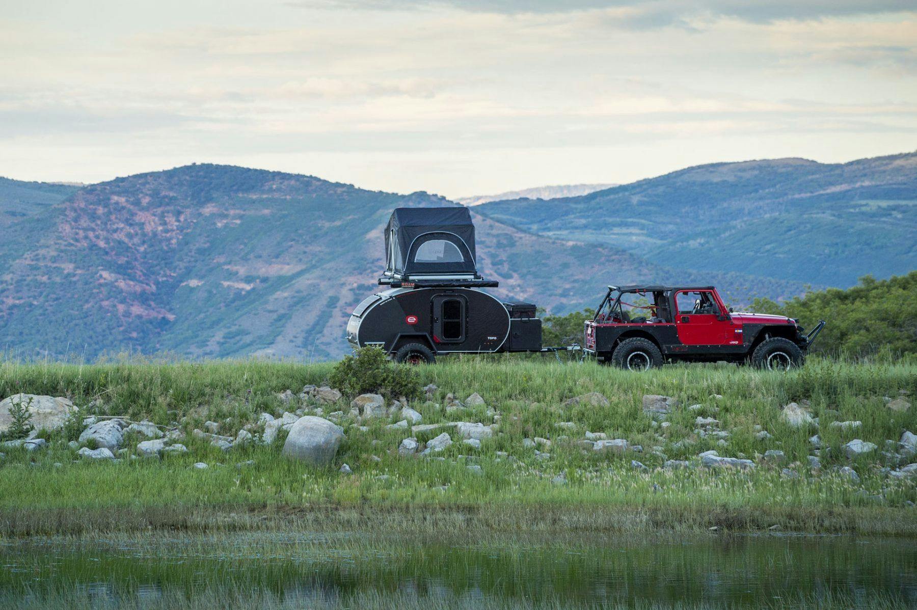 A teardrop trailer with a rooftop tent mounted on top being towed by a red JEEP.