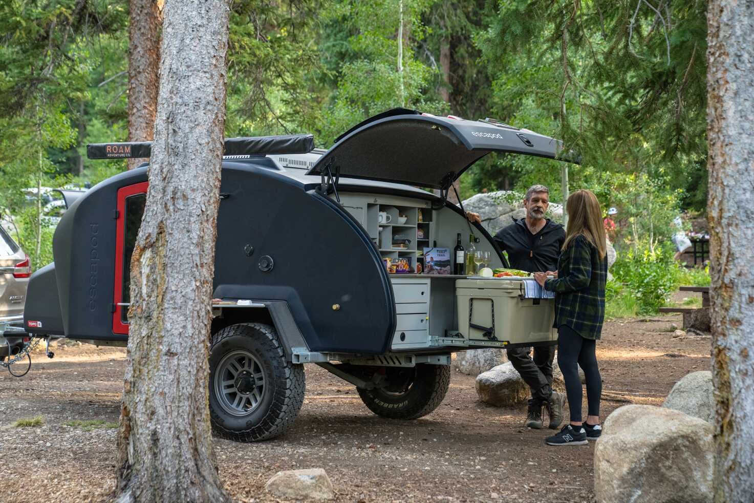 A middle aged couple is gathered around the kitchen galley of a TOPO2 teardrop camper enjoying some wine.