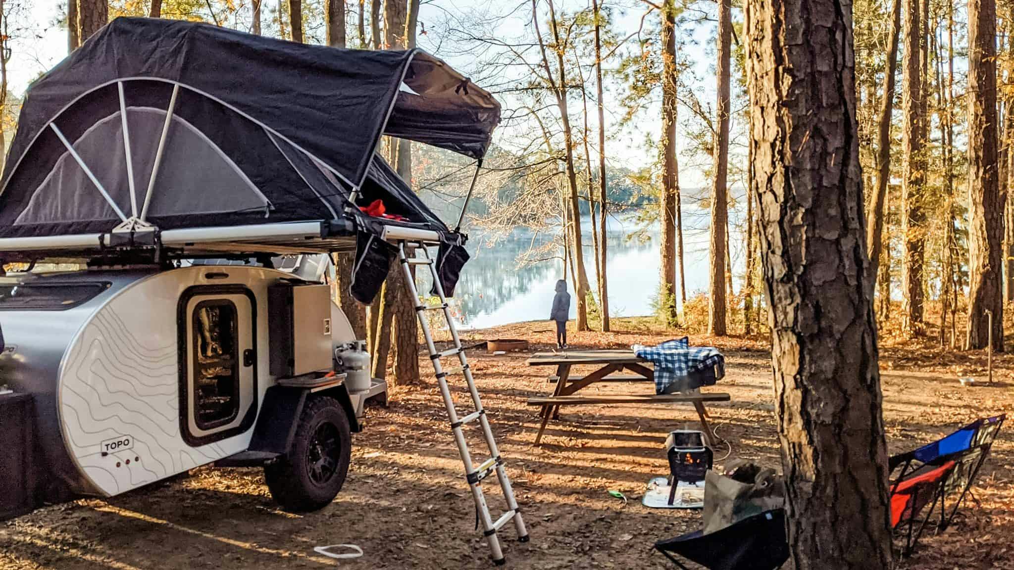 A teardrop camper with a rooftop tent deployed parked in near a lake and picnic table.