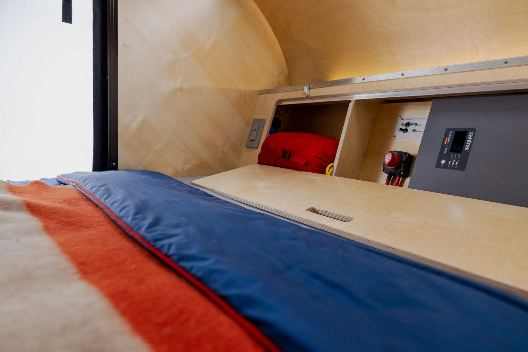 Birch Interior of a teardrop camper, with an angled headboard opened to show a Xantrex Inverter.
