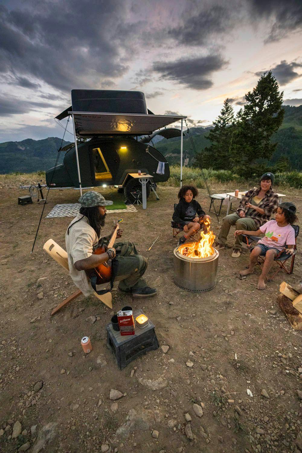 A family enjoying s'mores and music around a fire in front of their teardrop trailer.