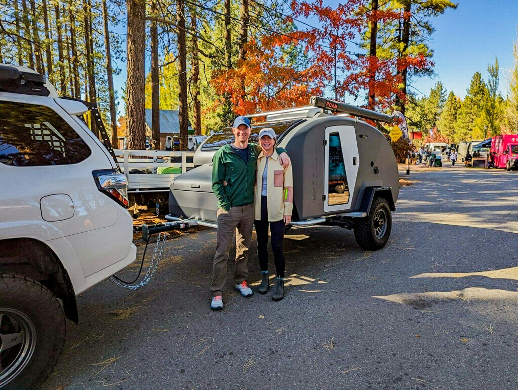 A couple stand in front of a Storm Grey TOPO2, a custom adventure teardrop trailer, by Escapod Trailers, in front of trees with fall leaves.