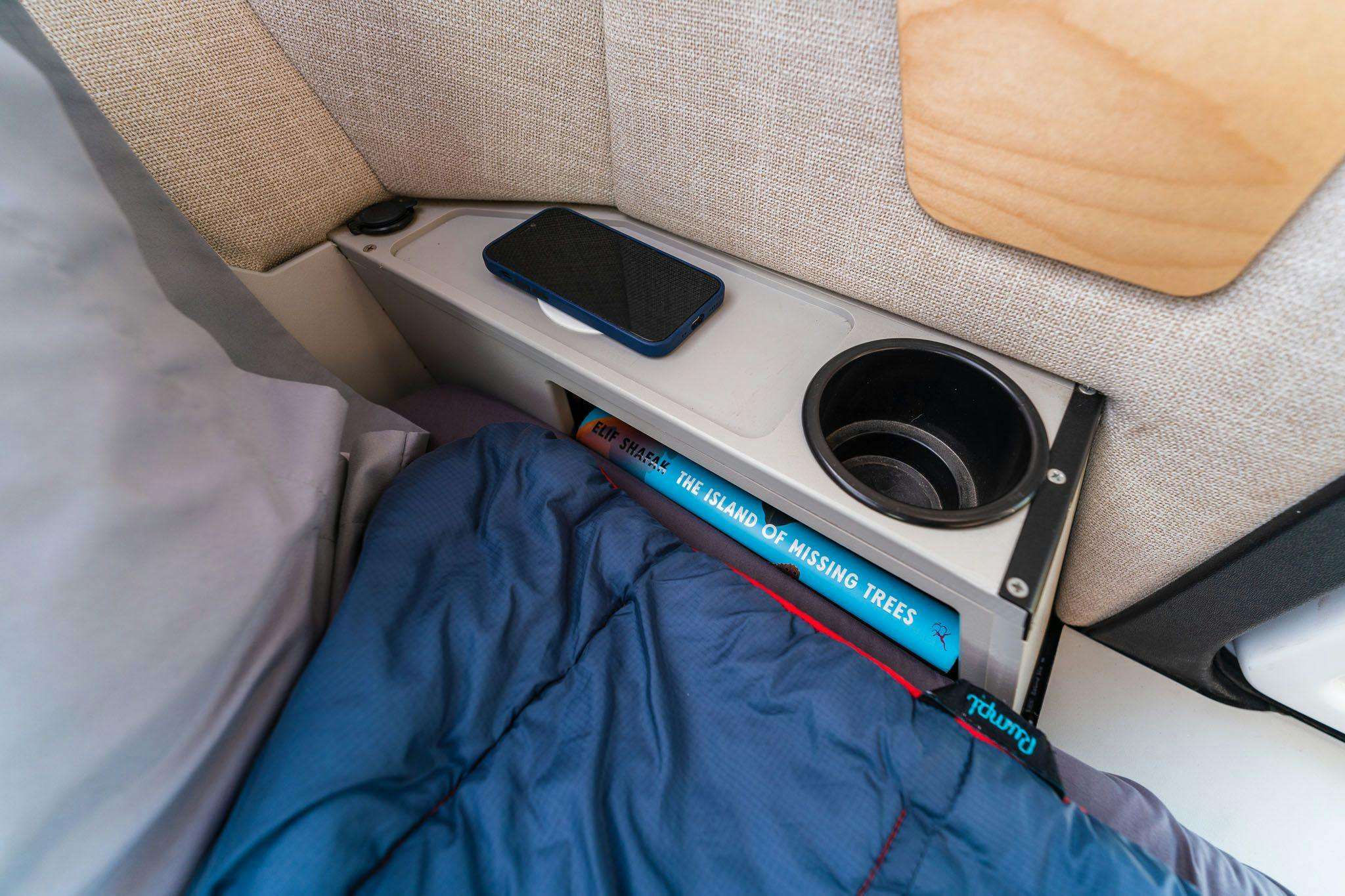 A book is peeking out of the lower storage of the nightstand in the TOPO2 teardrop trailer. There is a cell phone sitting on the wireless charging device.