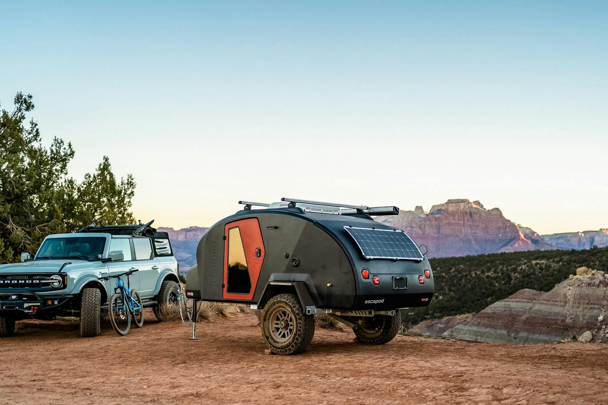 A navy blue teardrop camper being towed through desert landscape by a Ford Bronco.