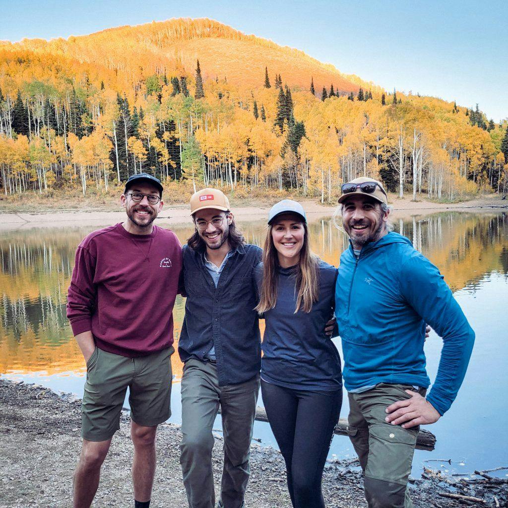 3 men and 1 woman standing infront of a lake with the fall colors in the background.