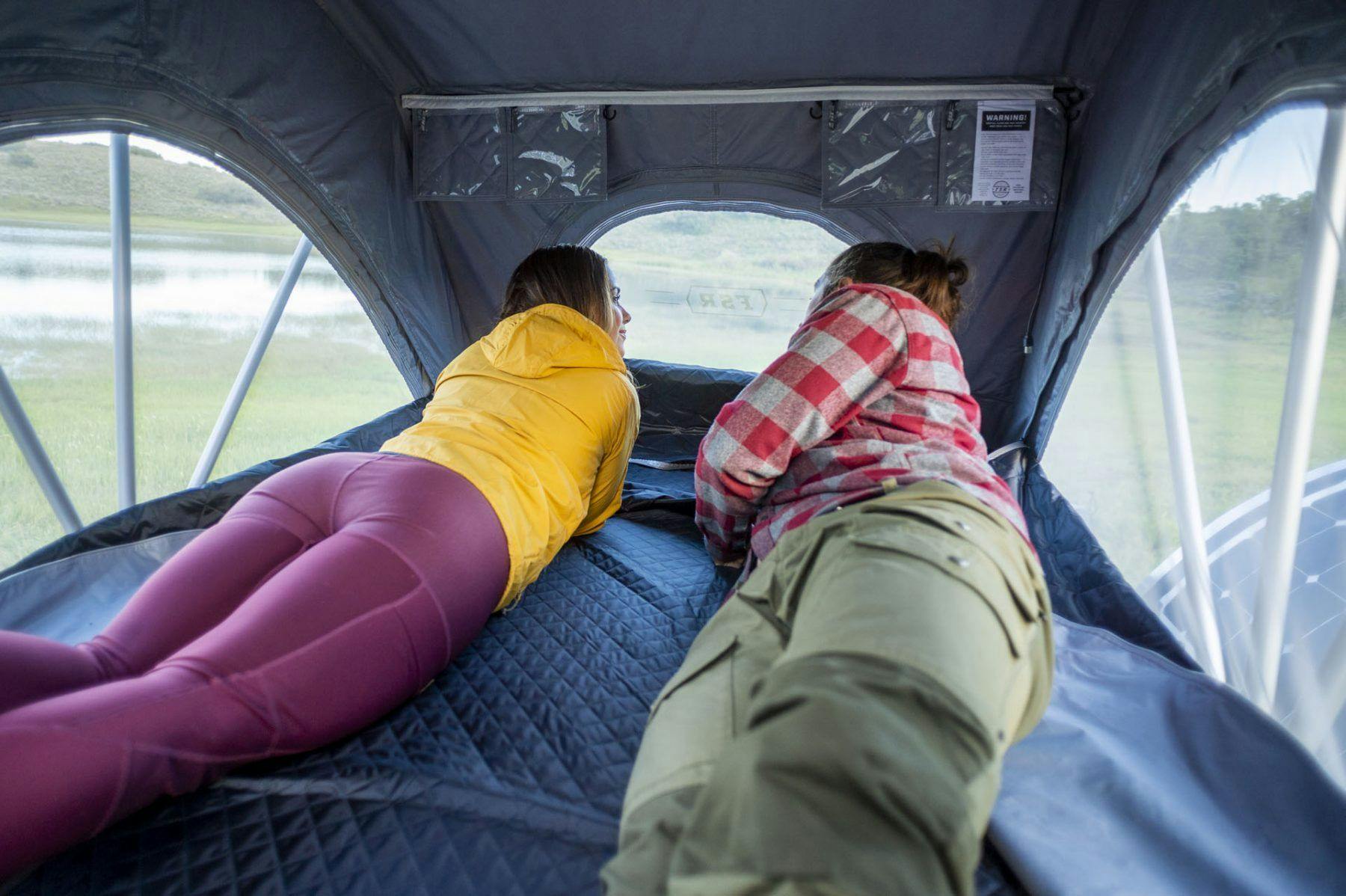 A couple laying in a rooftop tent mounted on top of a teardrop trailer.