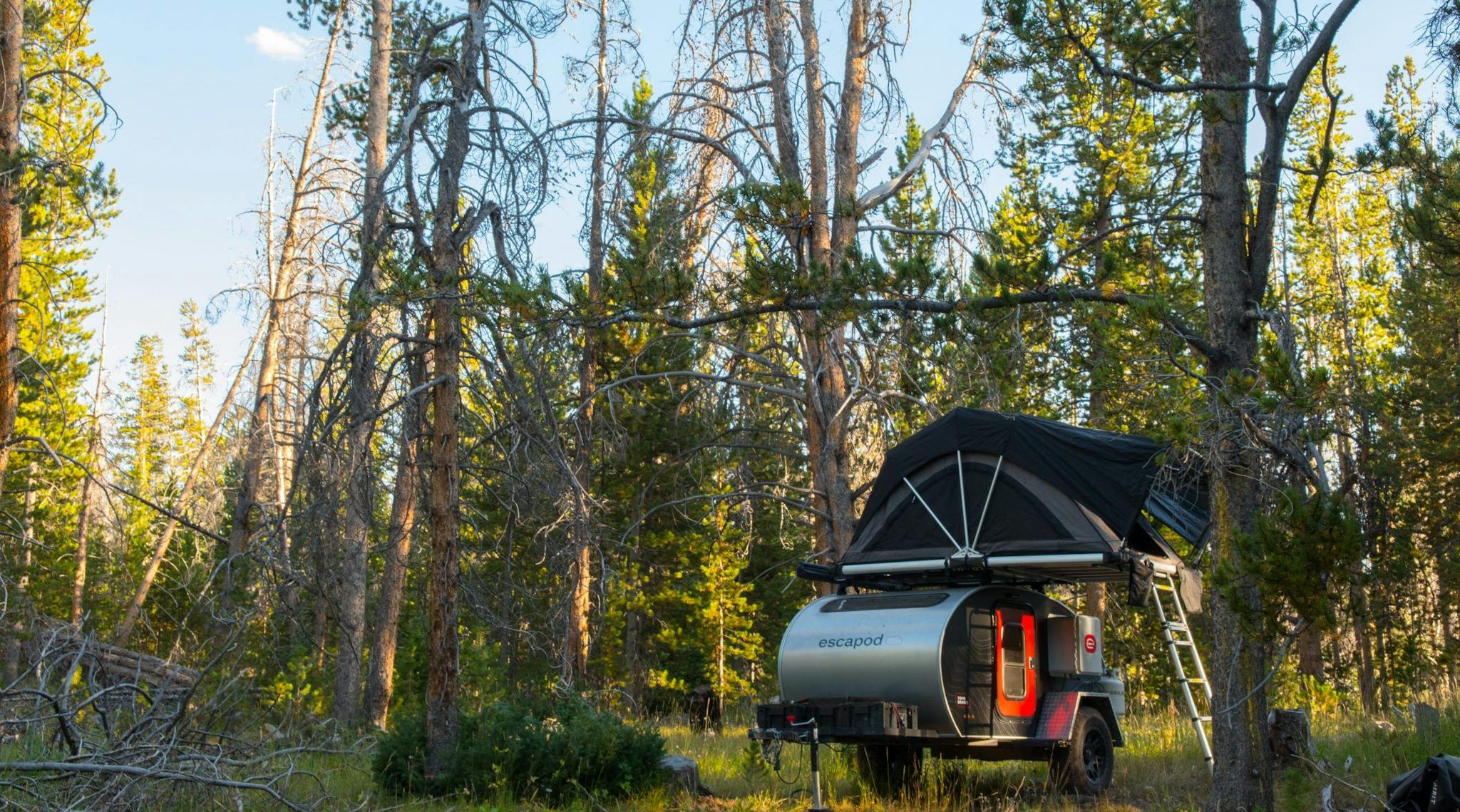 A black teardrop camper with a red door parked in a dispersed camping site with a rooftop tent mounted on top.