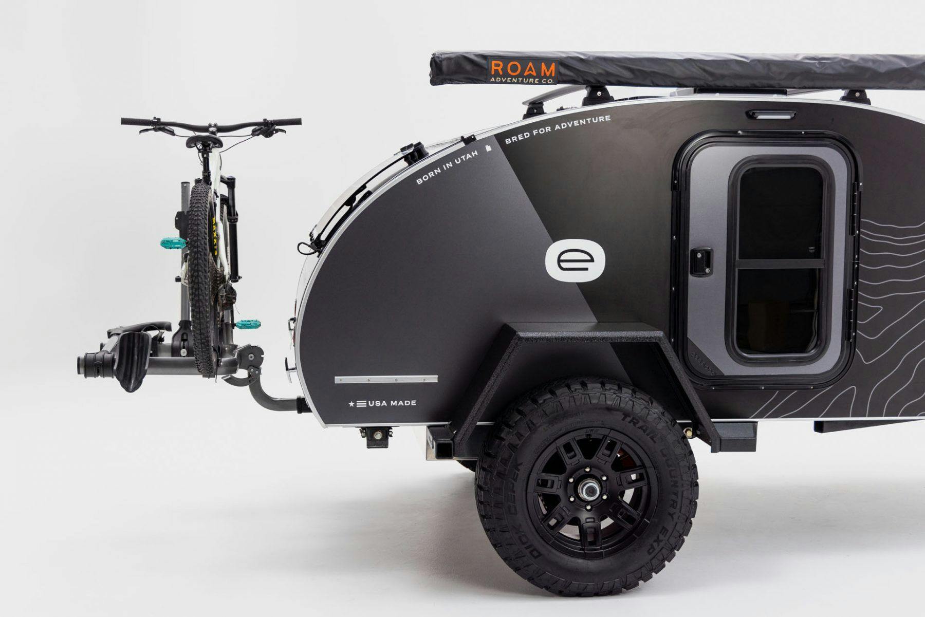 A profile view of an Escapod Original TOPO teardrop trailer is against an all-white backdrop with a mountain bike mounted on the rear hitch receiver.