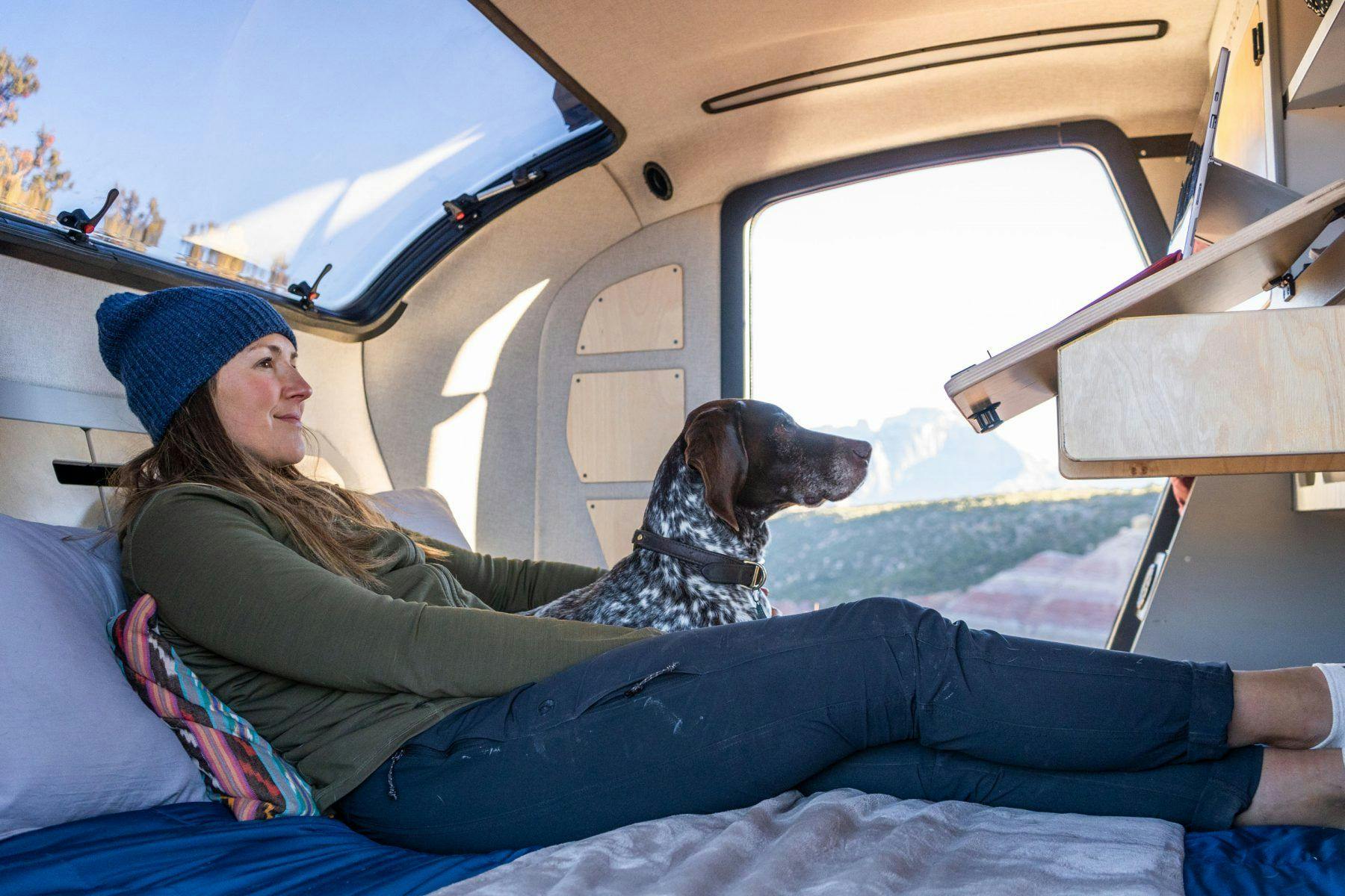 A woman and her dog cuddle in the cabin of a teardrop trailer. Comfortably relaxing and enjoying the scenery.