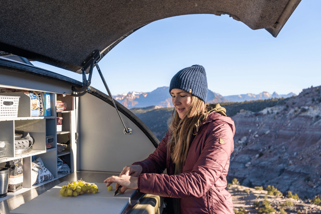 A woman in a purple jacket and blue beanie using the built in cutting board in the TOPO2, an offroad trailer, to prepare a meal.