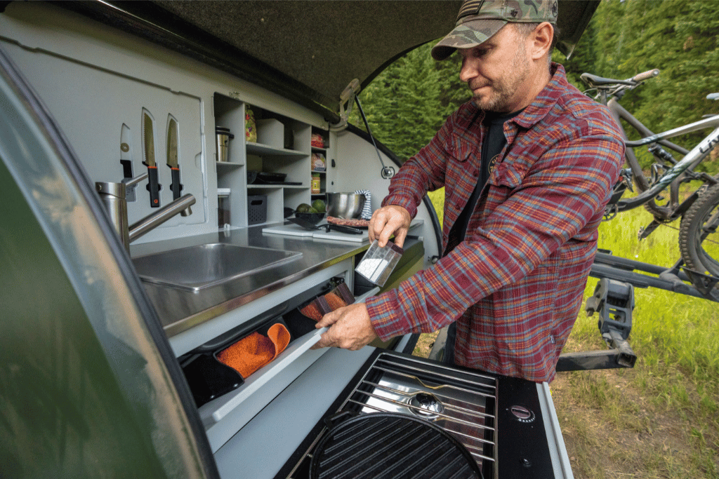 A man in a red plaid shirt is retreiving salt and pepper from the spice drawer in the galley of the Escapod TOPO2 teardrop camper.