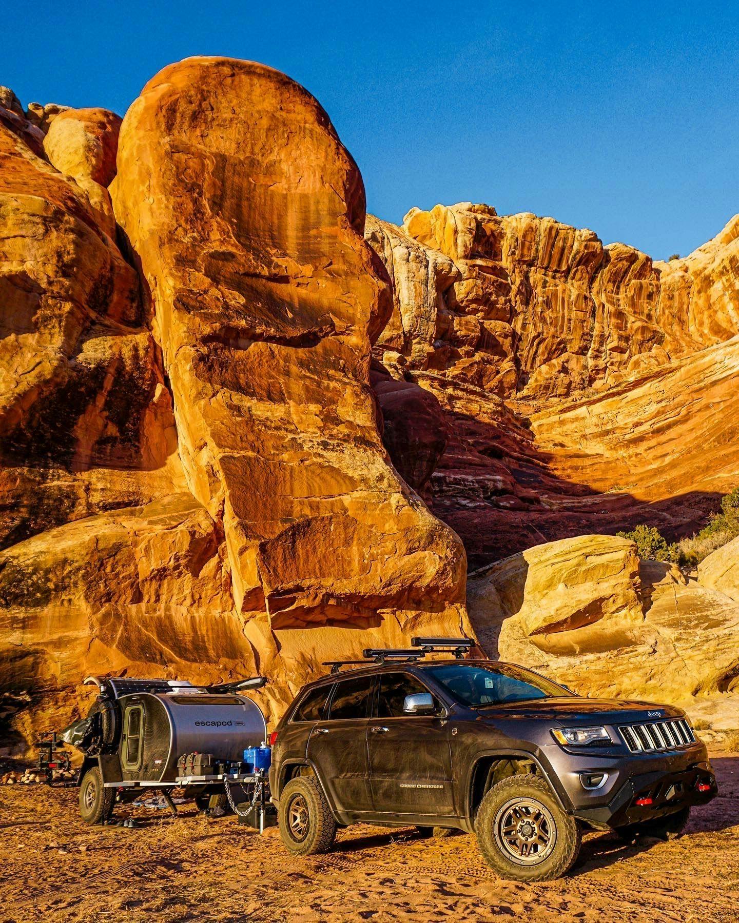 A Jeep towing an Escapod teardrop camper is nestled beneath glowing red rock in Thompson Springs, Utah.