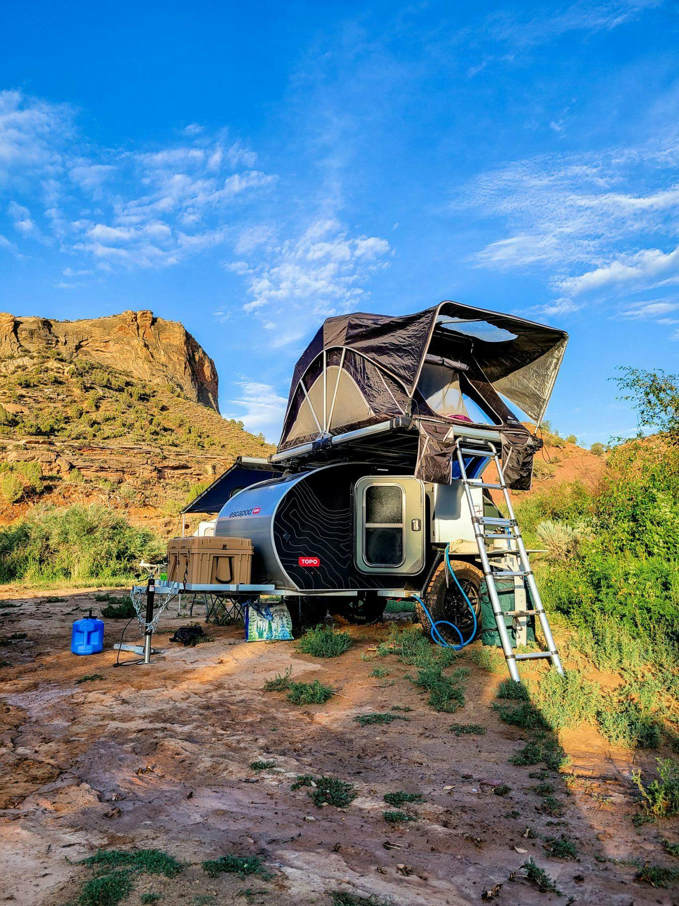 A black teardrop camper set up ina desert landscape with a rooftop tent mounted on top.