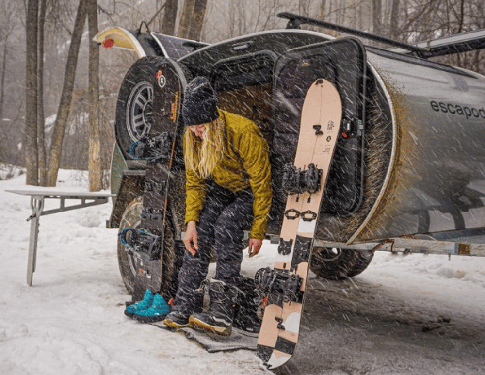 A woman changes into her snowboard boats in a snowstorm outside of her Original TOPO teardrop trailer by Escapod Trailers.