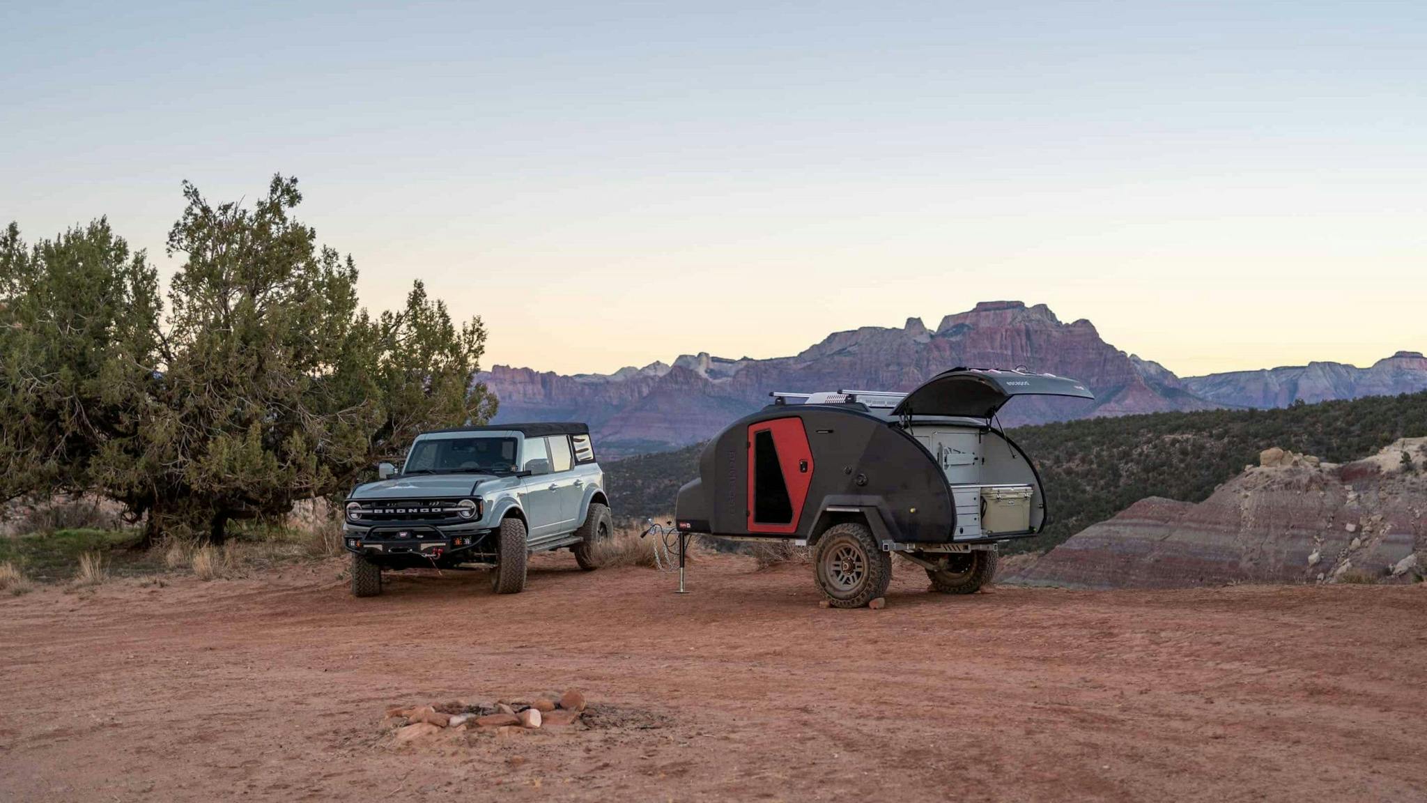 A navy blue teardrop camper being towed through desert landscape by a Ford Bronco.
