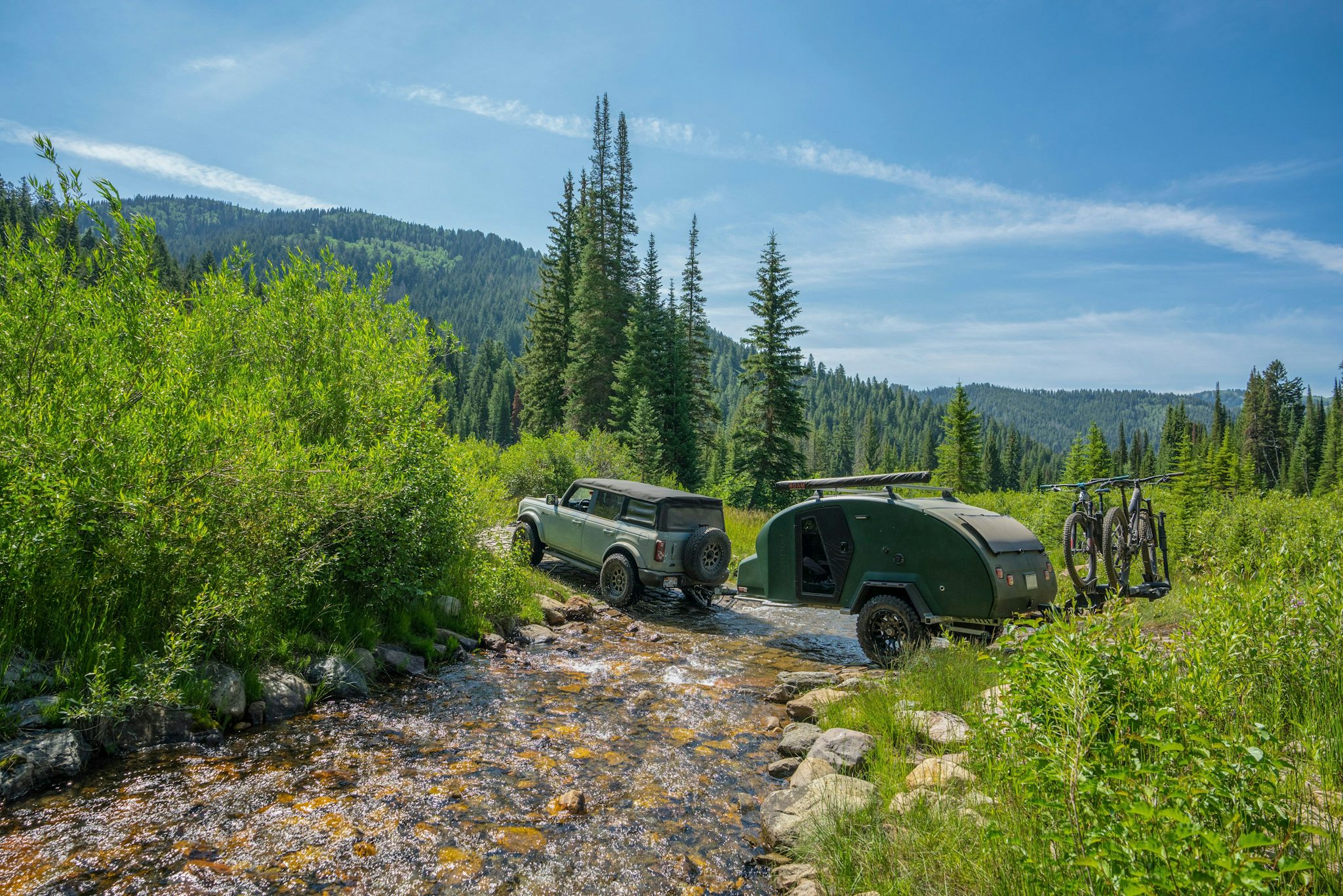 A Ford Bronco crosses a mountain stream while towing a TOPO2 off-road camping trailer. The grasses flanking the stream are bright green and tall pine trees cover the hillside in the distance.
