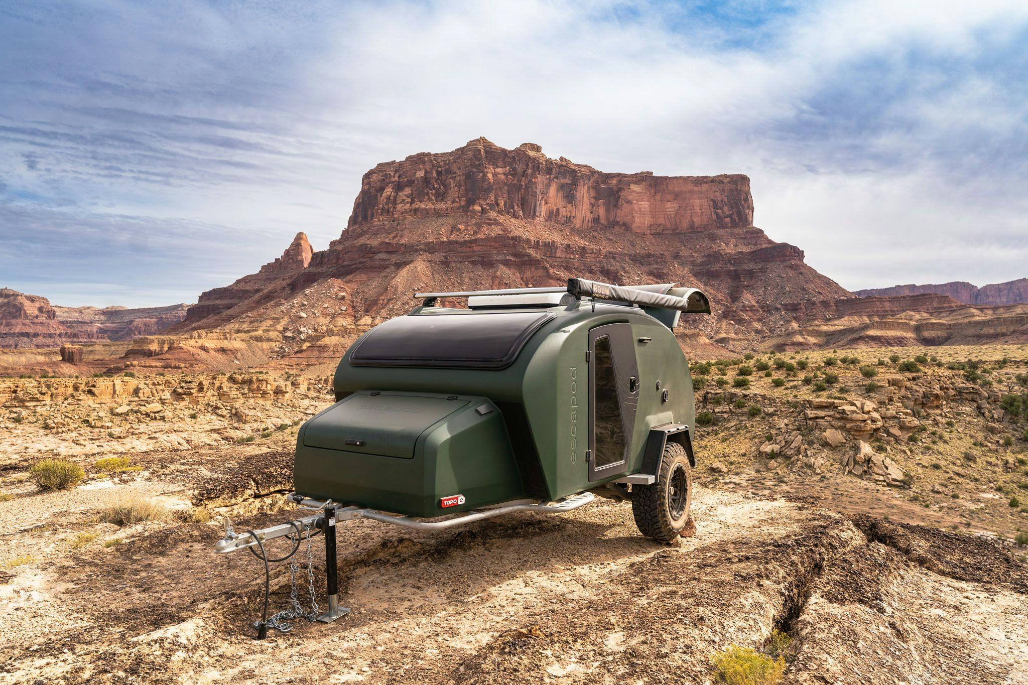 A green TOPO2 fiberglass camping trailer is parked on a rocky outcropping with a backdrop of a red rock mesa in the distance.