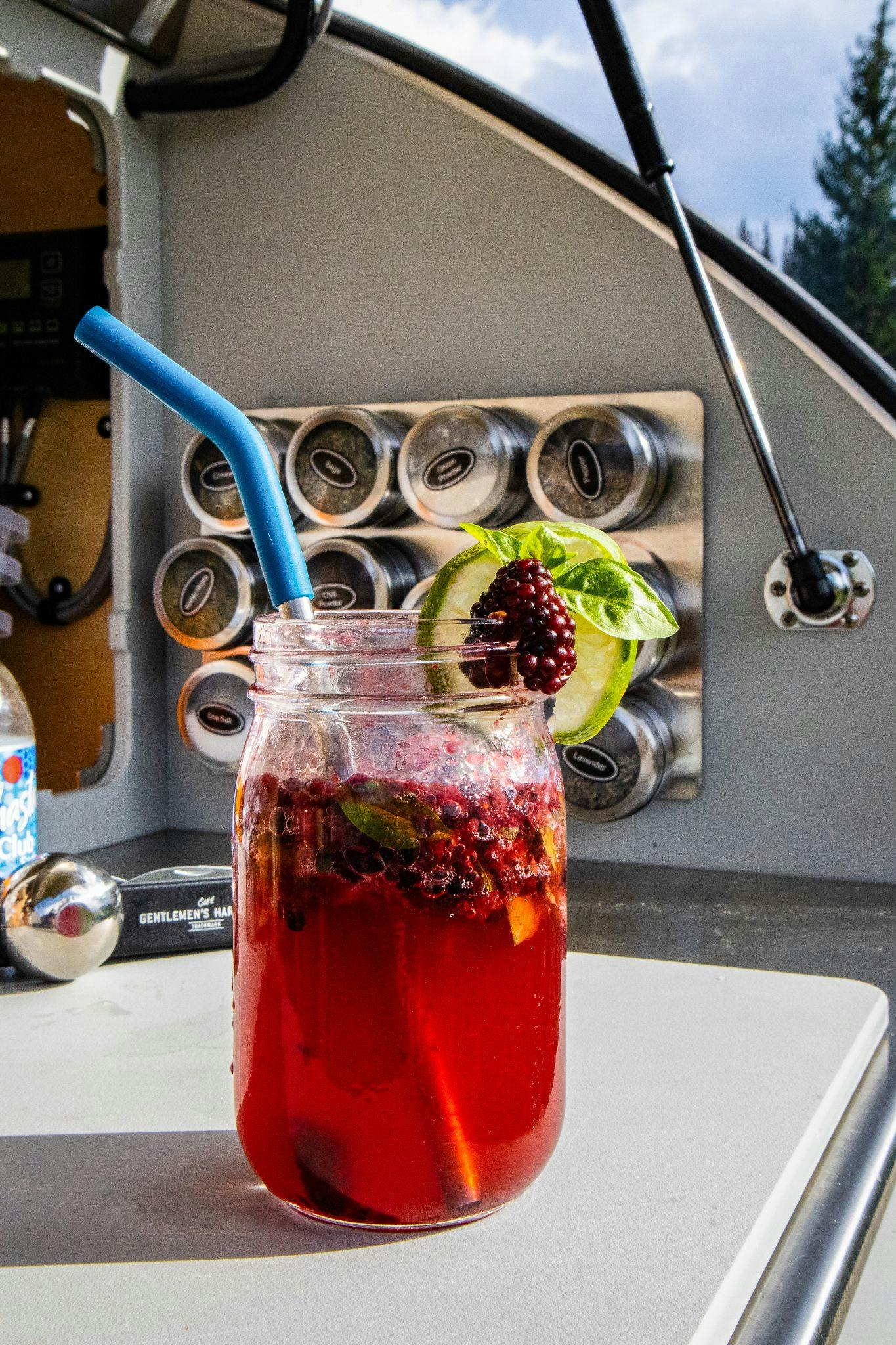 A red cocktail garnished with lime and blackberries set up in the galley of a teardrop camper.