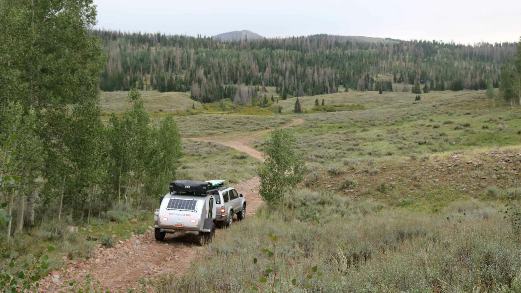 A teardrop camper being towed by a grey Toyota through a trail in the mountains.