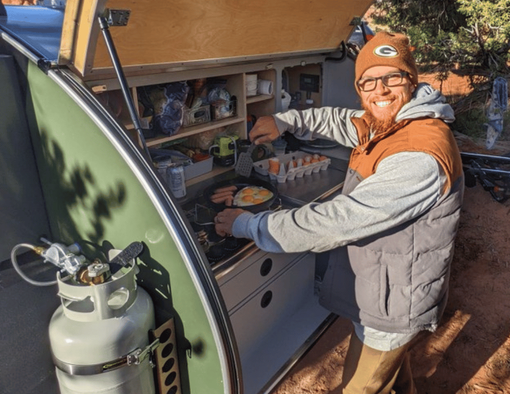A man smiles at the camera as he is cooking a breakfast meal in the galley of his Original TOPO trailer, a teardrop trailer by Escapod Trailers.