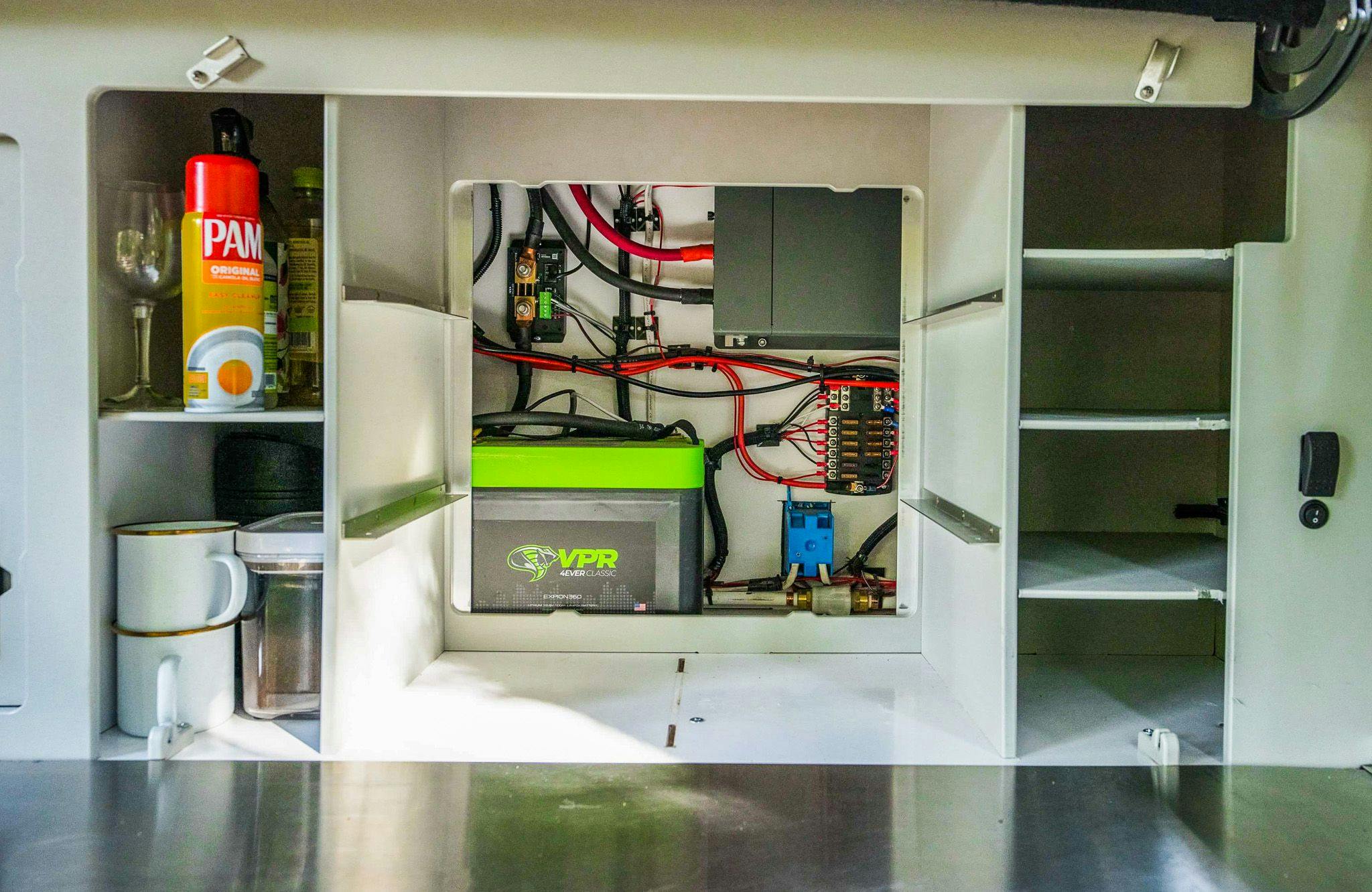 Behind the modular shelving in a teardrop trailer galley, shows the interior mechanicals. This includes a battery setup to power the trailer.