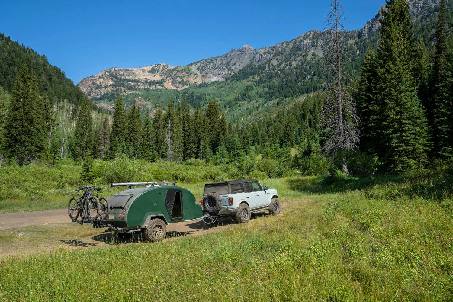 A teardrop trailer being towed by a Ford Bronco, arriving at a beatuiful campsite surround by pine trees, green grass, and mountains.