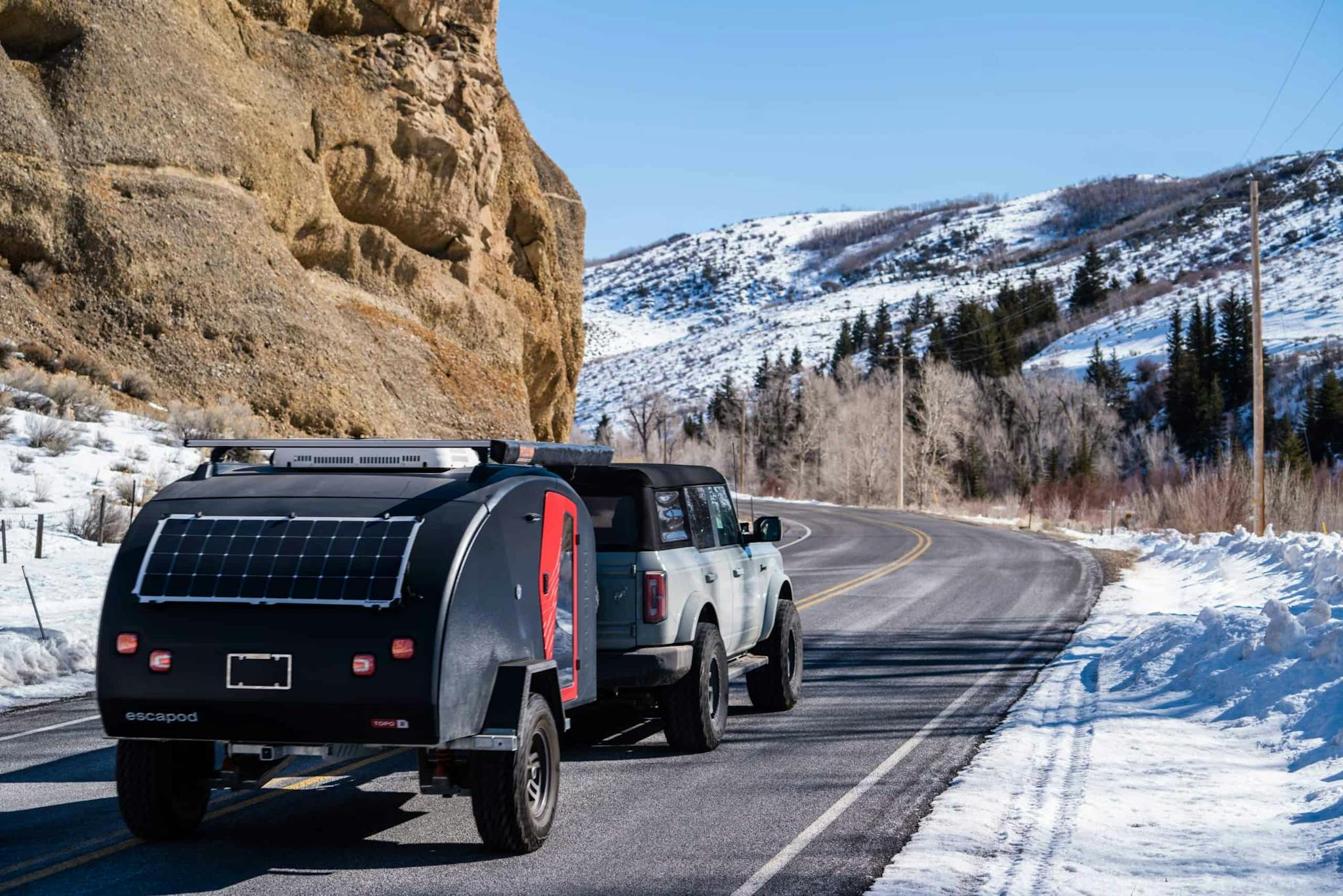 A Bronco is towing a navy blue teardrop camper with a red door down a paved snowy road.