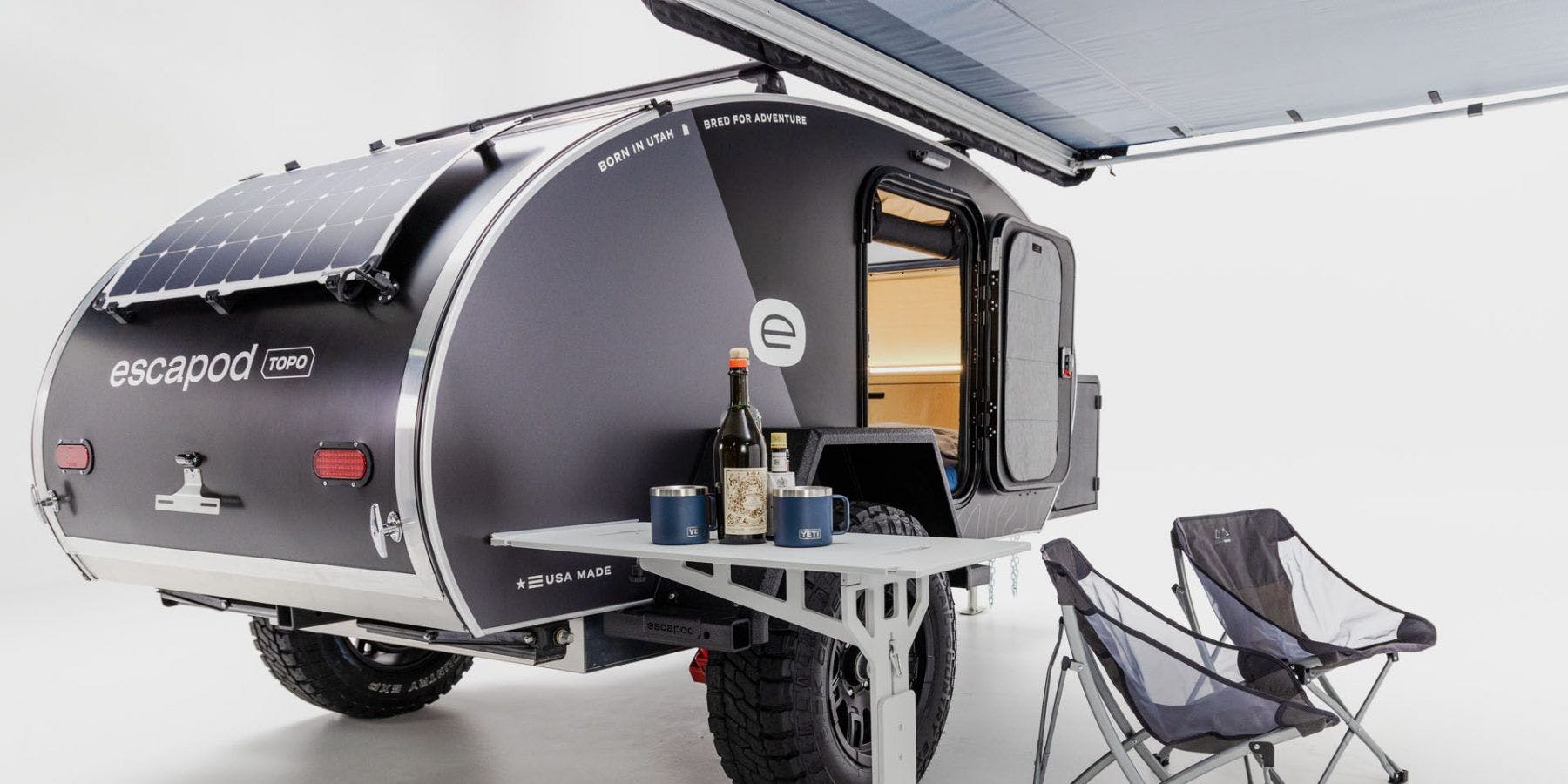 An Escapod Original TOPO teardrop camper in a studio against a white backdrop. A solar panel and spare tire accent the trailer.