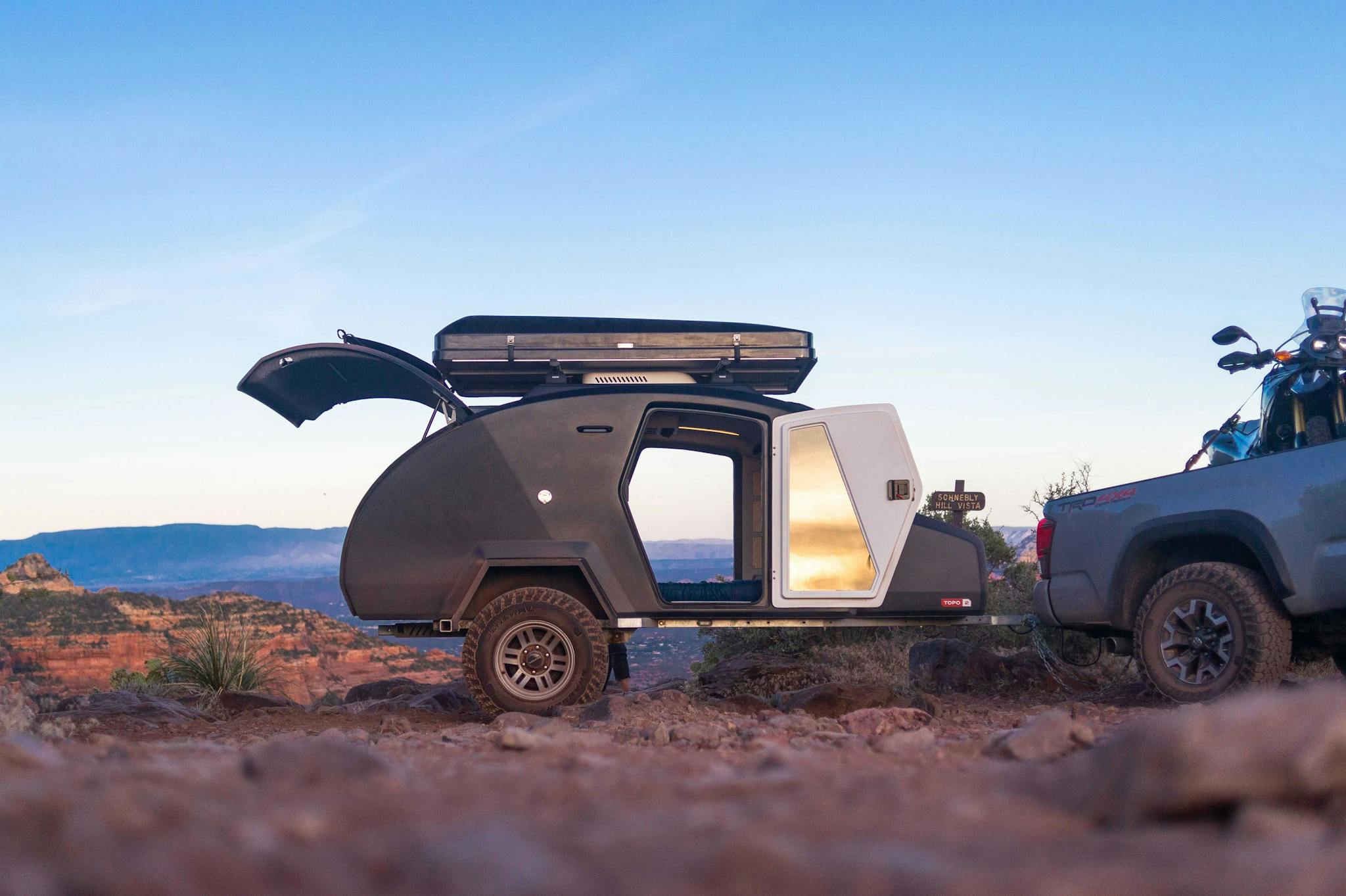An Escapod TOPO2 teardrop camper is sitting on the edge of a desert cliff illuminated by the rising sun.