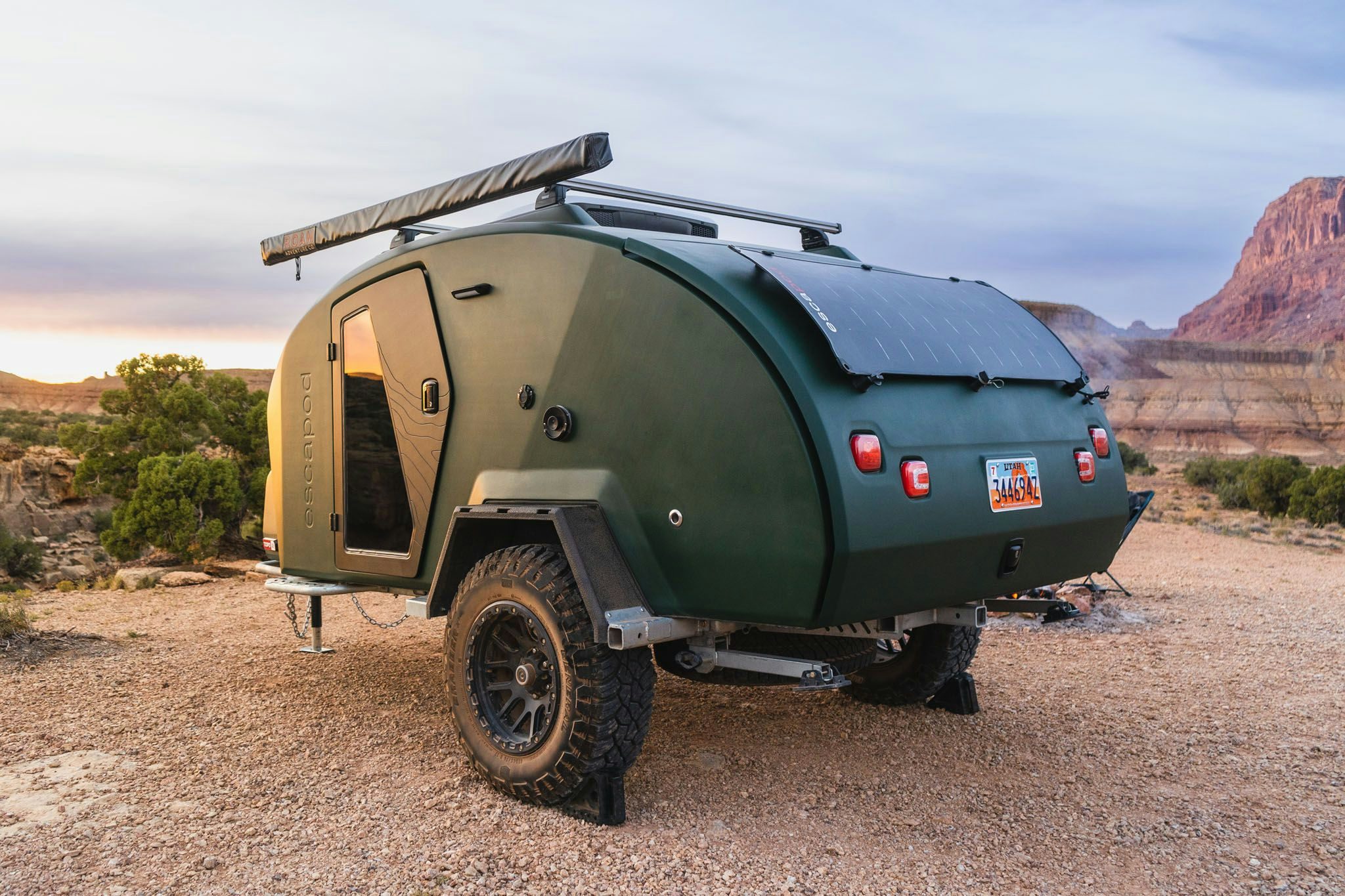 A green teardrop trailer parked in a desert landscape at sunset.
