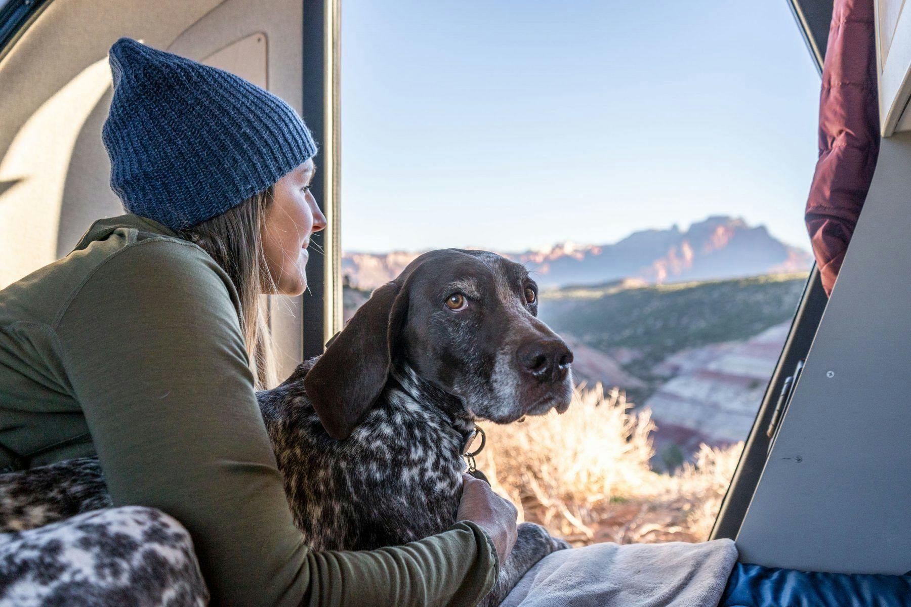 A woman wearing a green jacket and a blue beanie and her dog enjoying the cabin of a teardrop camper.