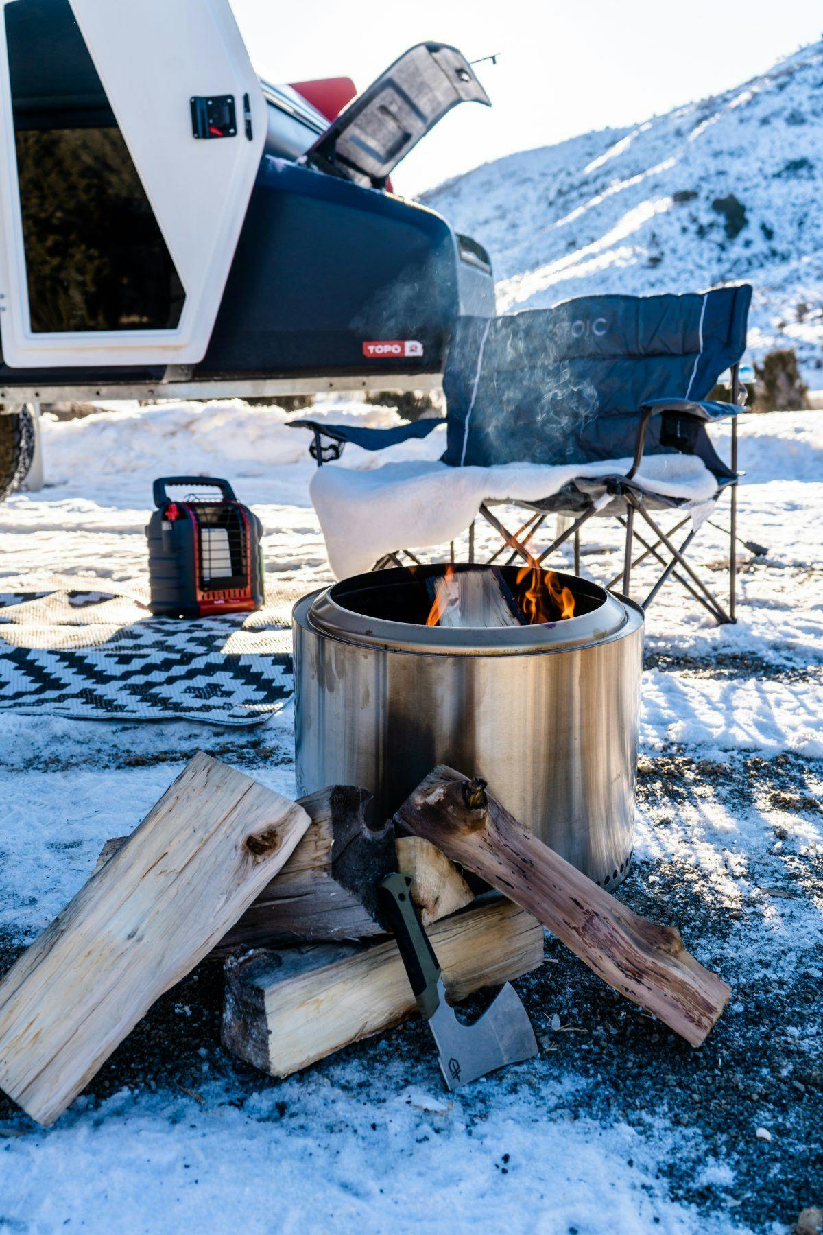 A SOLO stove set up at camp near a teardrop trailer in a winter wonderland.