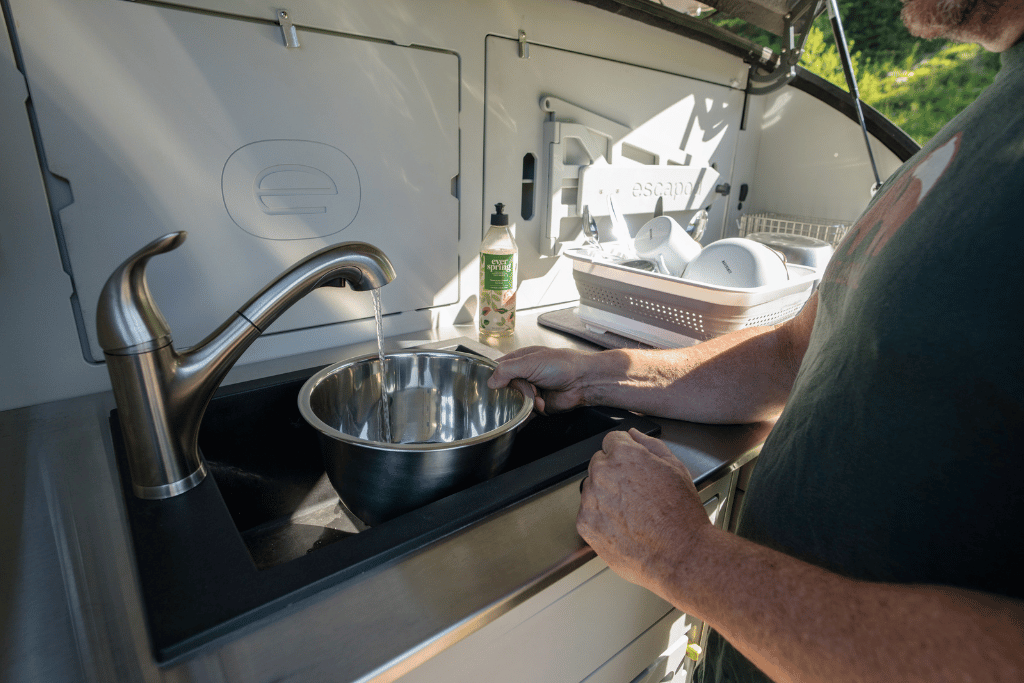 A man is a green shirt is using the full sized sink in the TOPO2, an offroad trailer, to wash the dishes while camping.