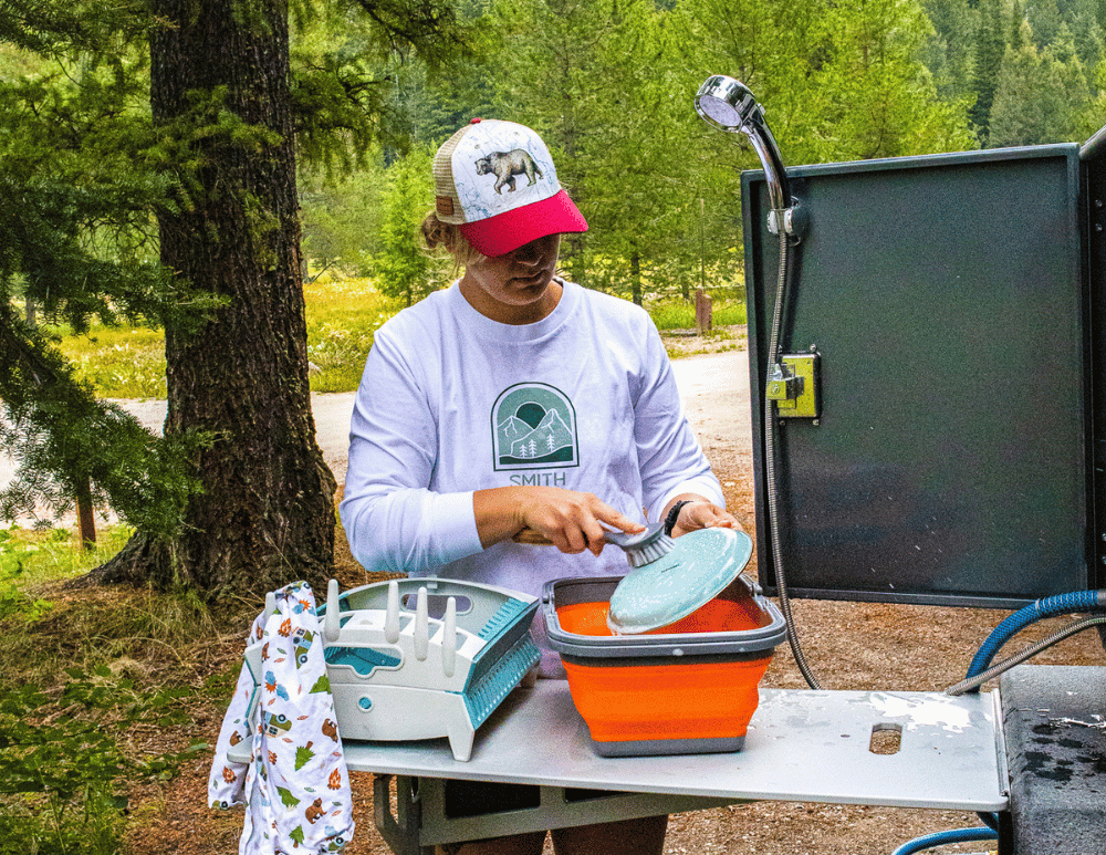 A woman in a baseball cap and long sleeved shirt, washing dishes outside of her Original TOPO teardrop trailer, by Escapod Trailers.