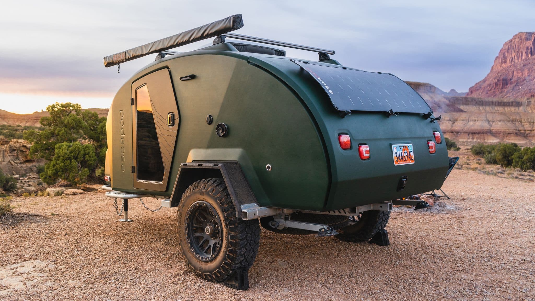 A green Escapod TOPO2 trailer with a solar panel mounted on the rear, parked at a rugged desert campsite.