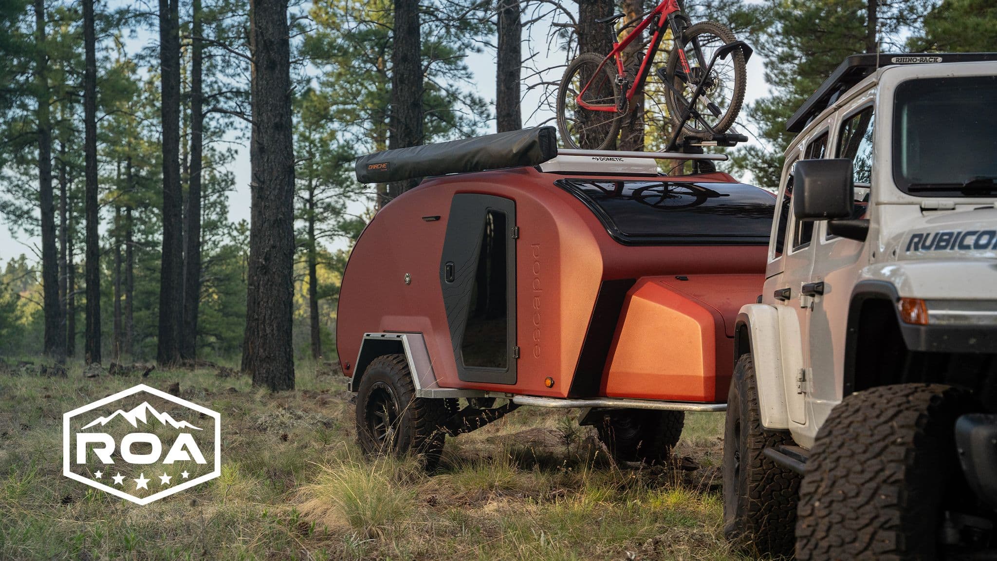 A Jeep Rubicon tows an Escapod TOPO2 trailer in a pine forest, with a mountain bike mounted on top and an "ROA" logo in the corner.