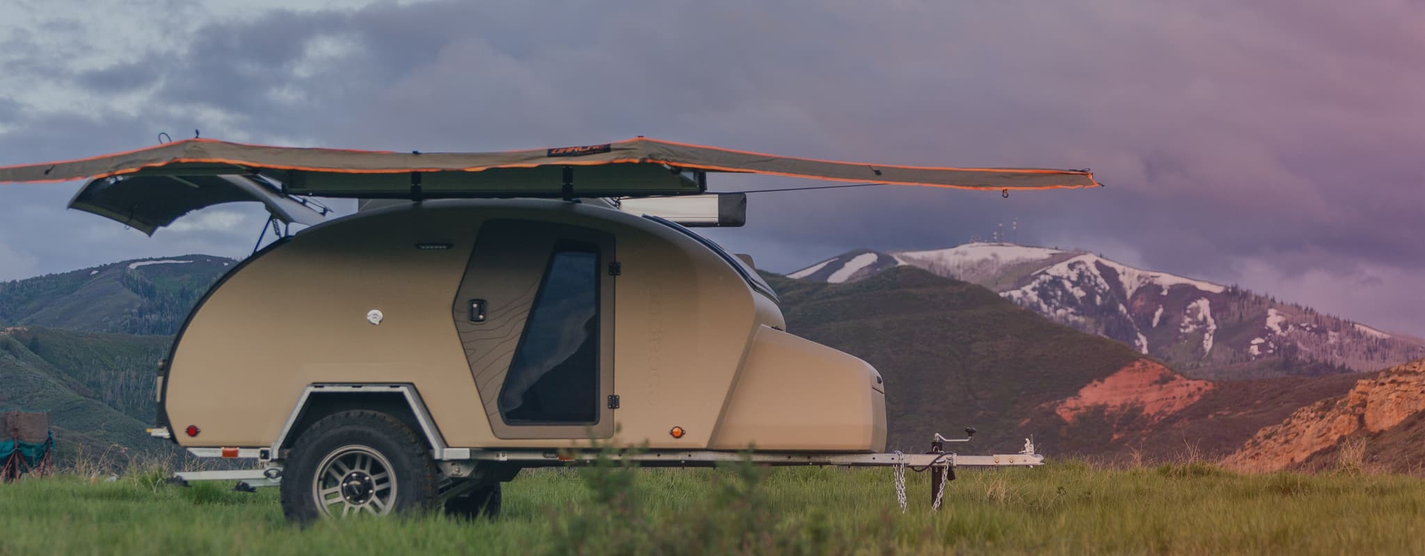 Side profile of an Escapod TOPO2 trailer parked on a grassy hill, equipped with an awning, with snow-capped mountains and a sunset sky in the background.