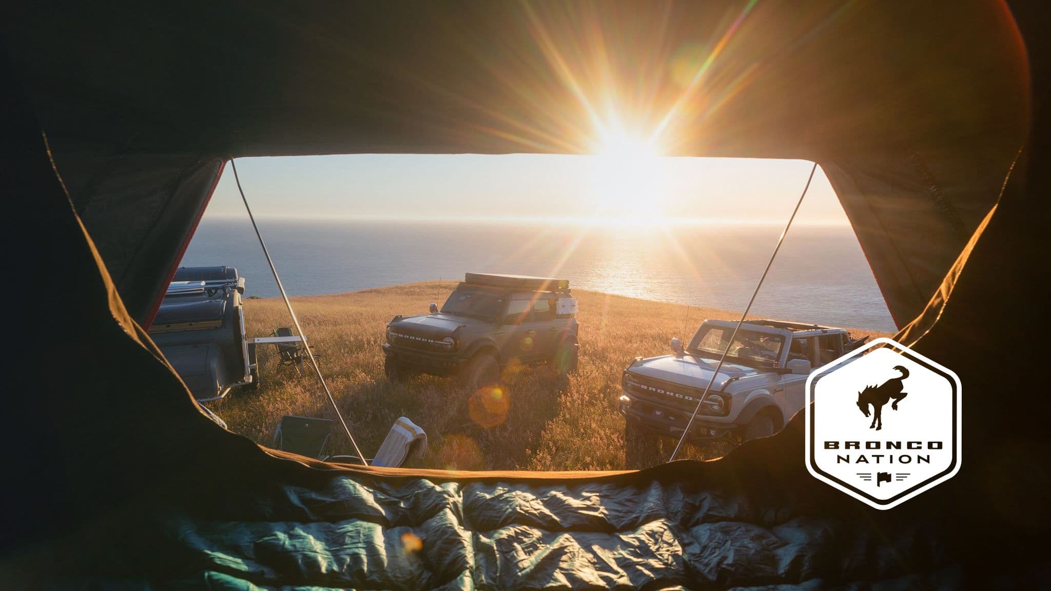 View from inside a tent overlooking Ford Broncos parked on a grassy coastal bluff at sunset, with a "Bronco Nation" logo in the corner.