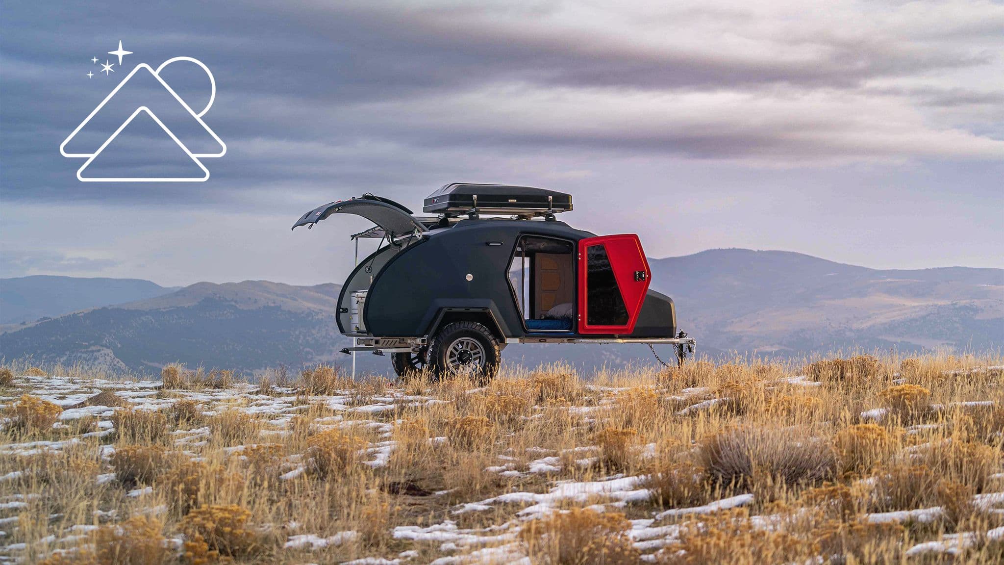 An Escapod TOPO2 off-road trailer with a red door sits on a rugged, snowy plateau with a mountainous landscape under a cloudy sky.