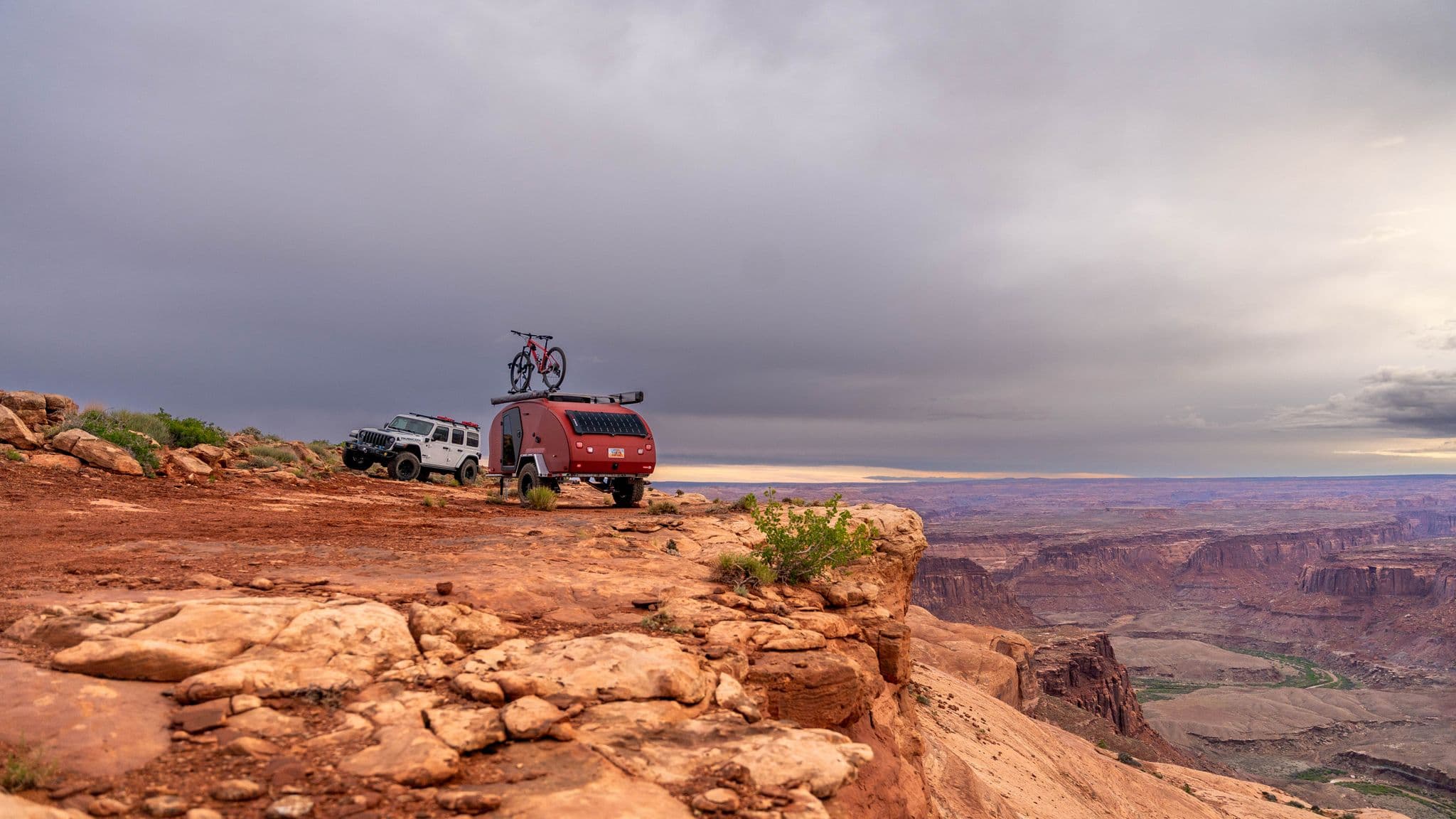 A Jeep Wrangler and an Escapod TOPO2 trailer with a mountain bike on top are parked on a rocky cliffside overlooking a vast canyon under a cloudy sky.
