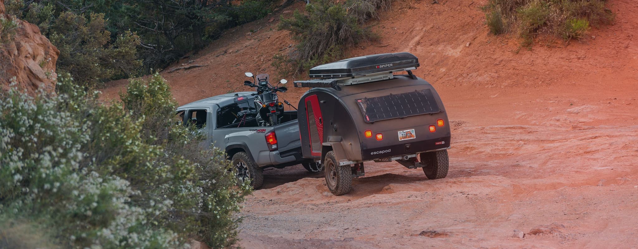 Rear view of an Escapod TOPO2 trailer hitched to a Tacoma truck with a dirt bike in the bed, navigating a steep, rocky desert trail.