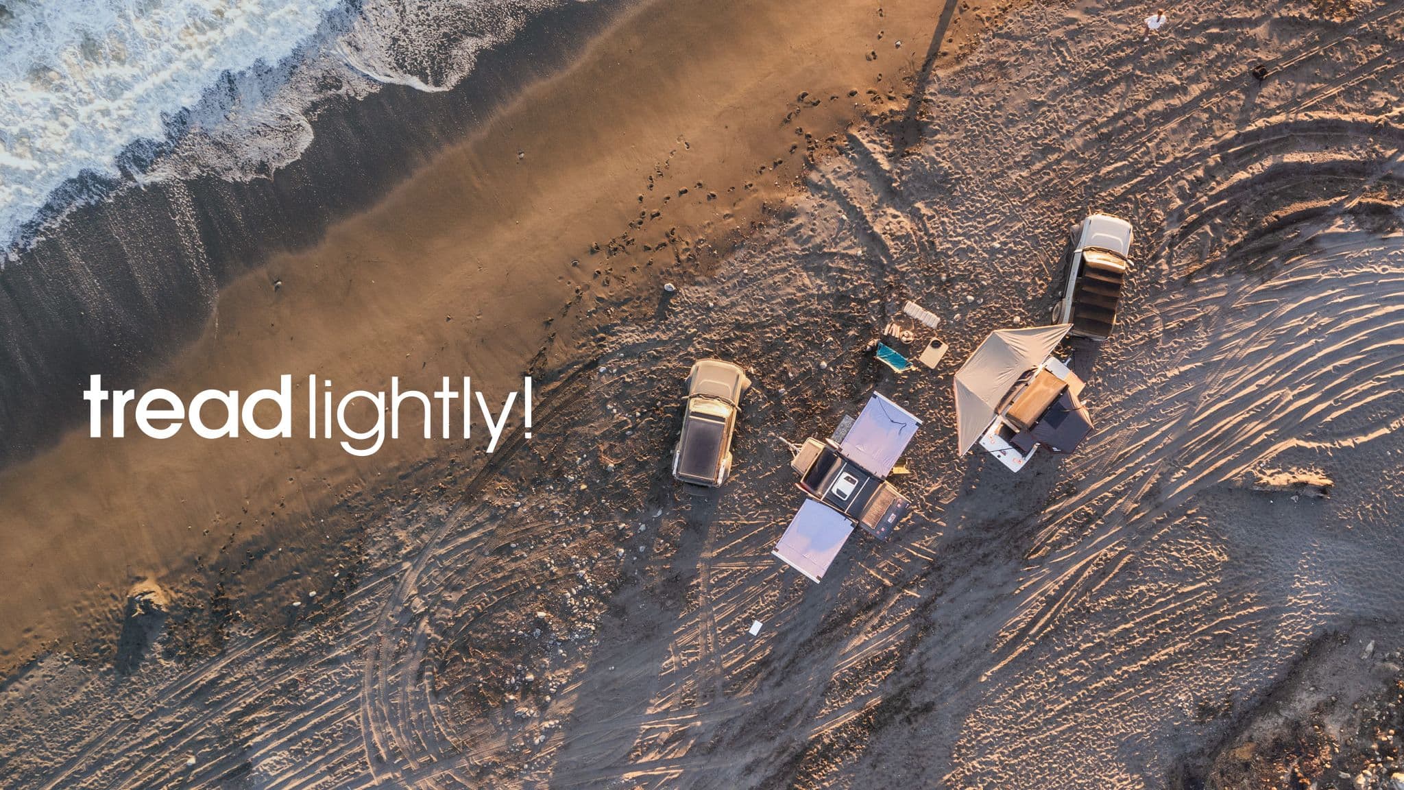Aerial view of an off-road beach campsite with trucks and tents near the shoreline, featuring the text "tread lightly!" in the sand.
